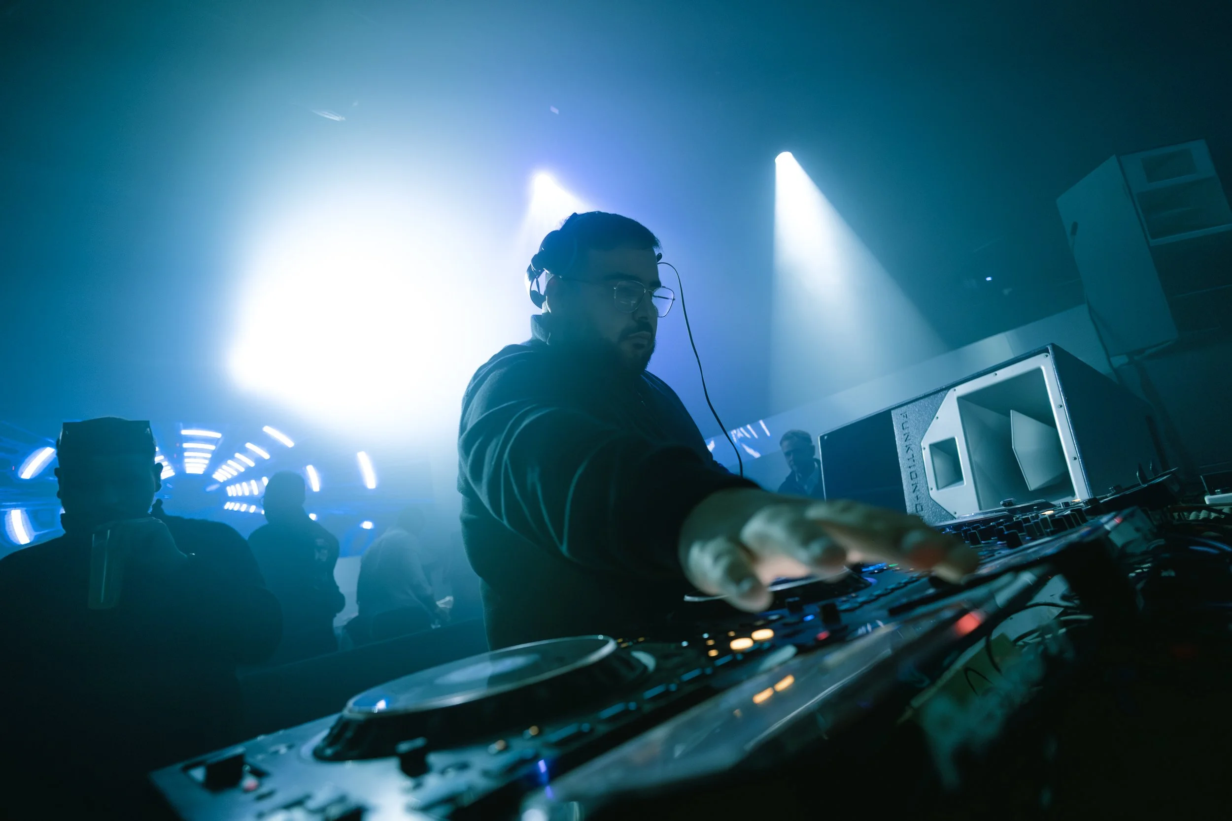 DJ wearing headphones playing music at a nightclub with blue lighting and silhouettes of people dancing in the background.