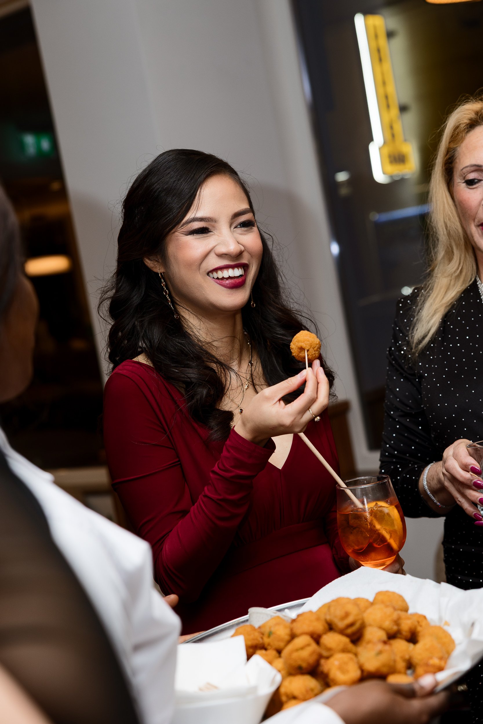 A woman in a red dress smiling while holding a skewer with breaded fried food, with a drink and a tray of fried food on the table.
