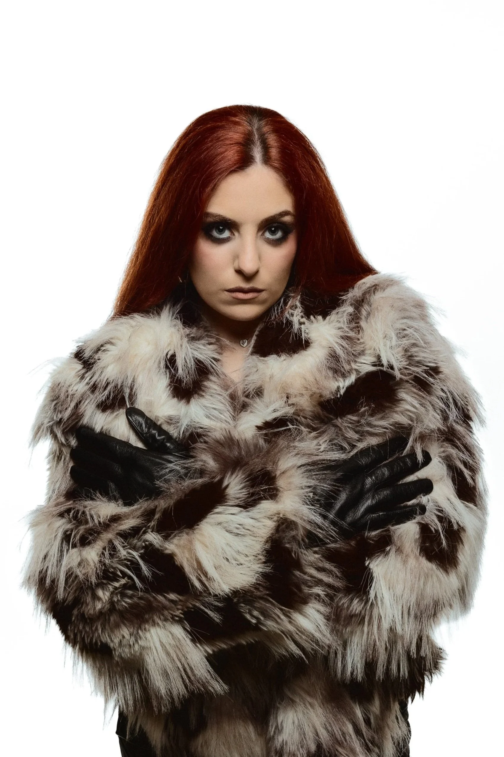 A woman with red hair and dark eye makeup wearing a fur coat and black gloves, looking directly at the camera against a plain white background.