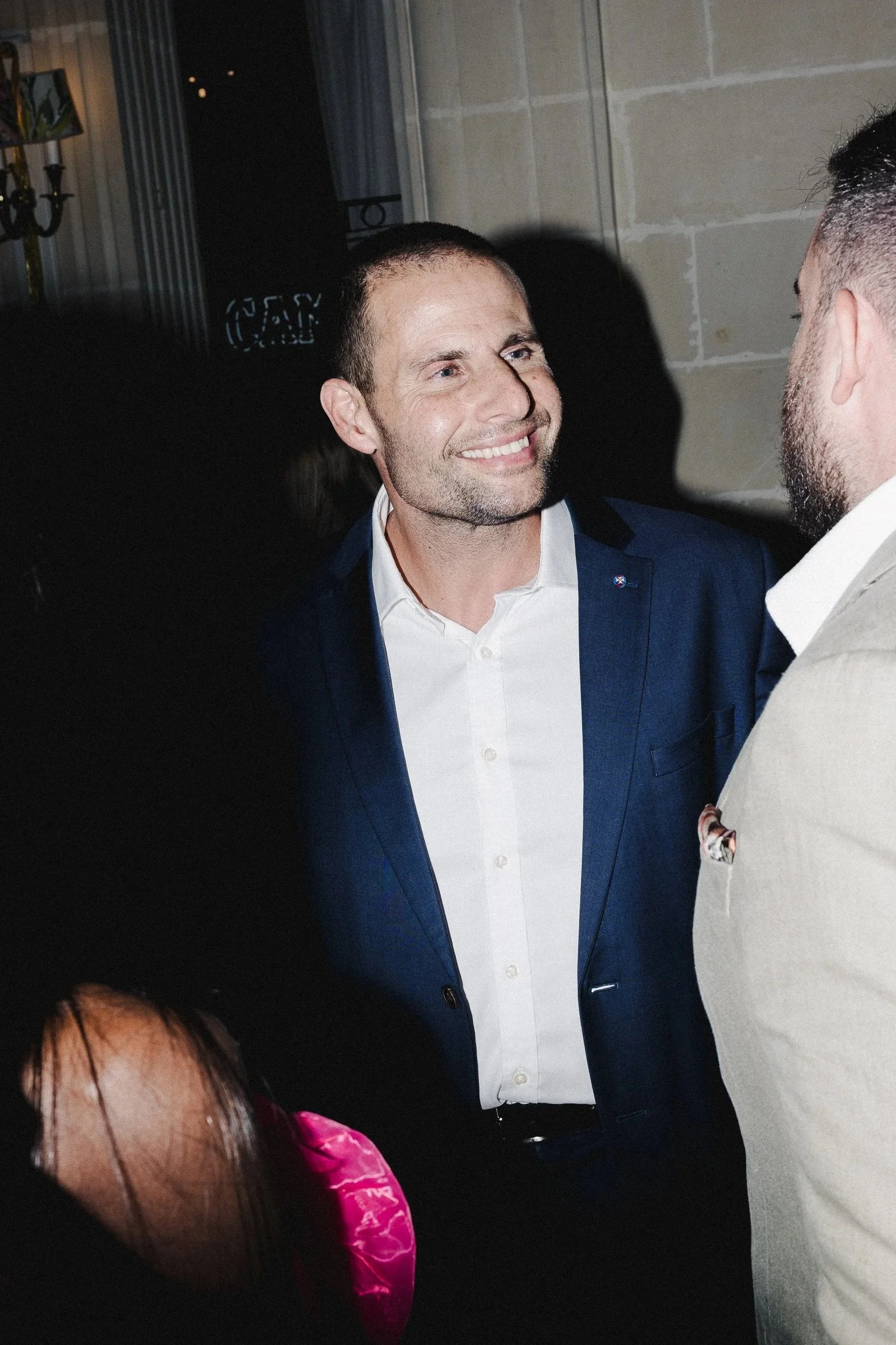 A man in a dark suit smiling and talking to another man in a beige suit at a social event.