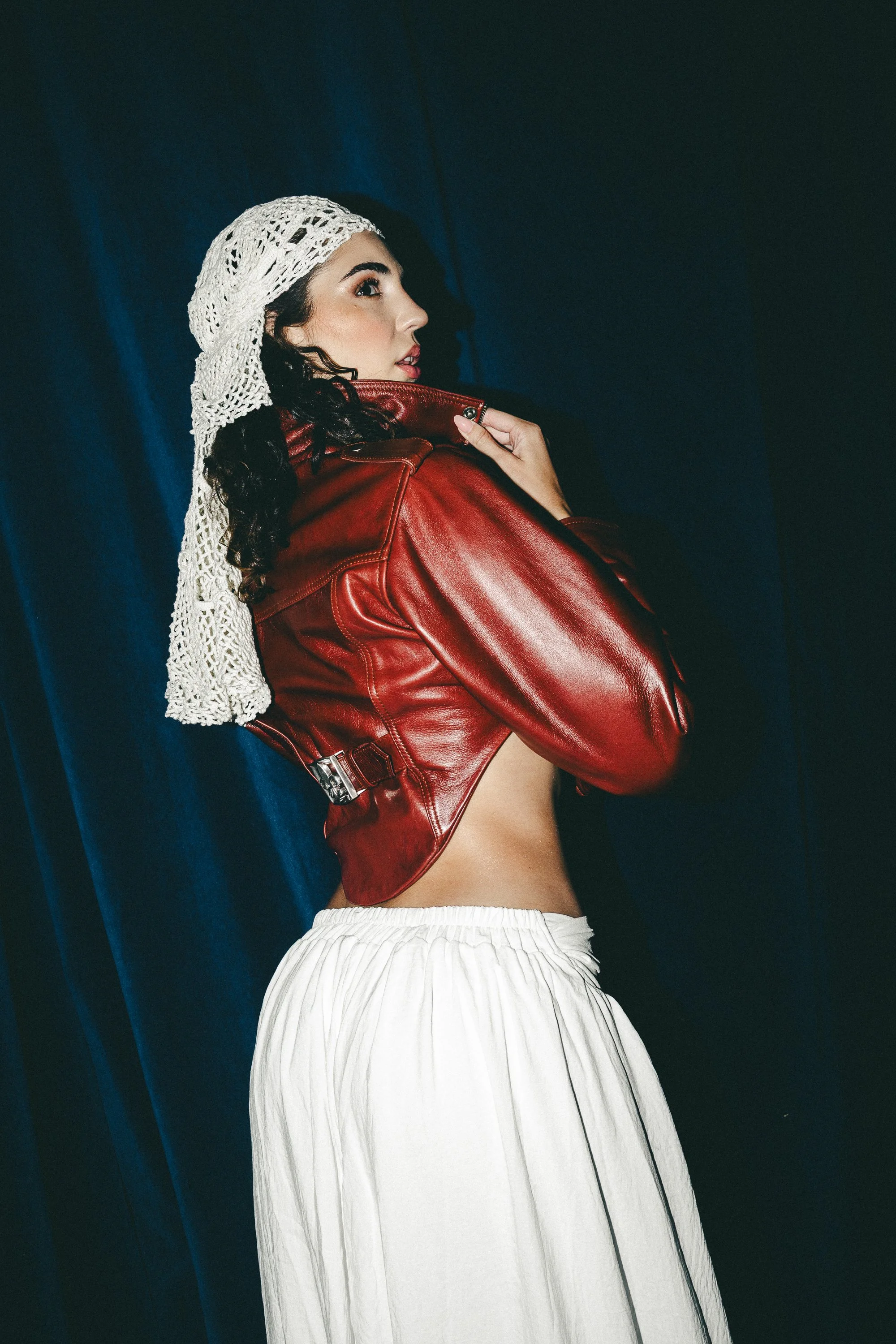 A woman with dark, curly hair and bold makeup wearing a white knitted hat, a red leather jacket, and white flowy pants standing against a dark blue curtain background.