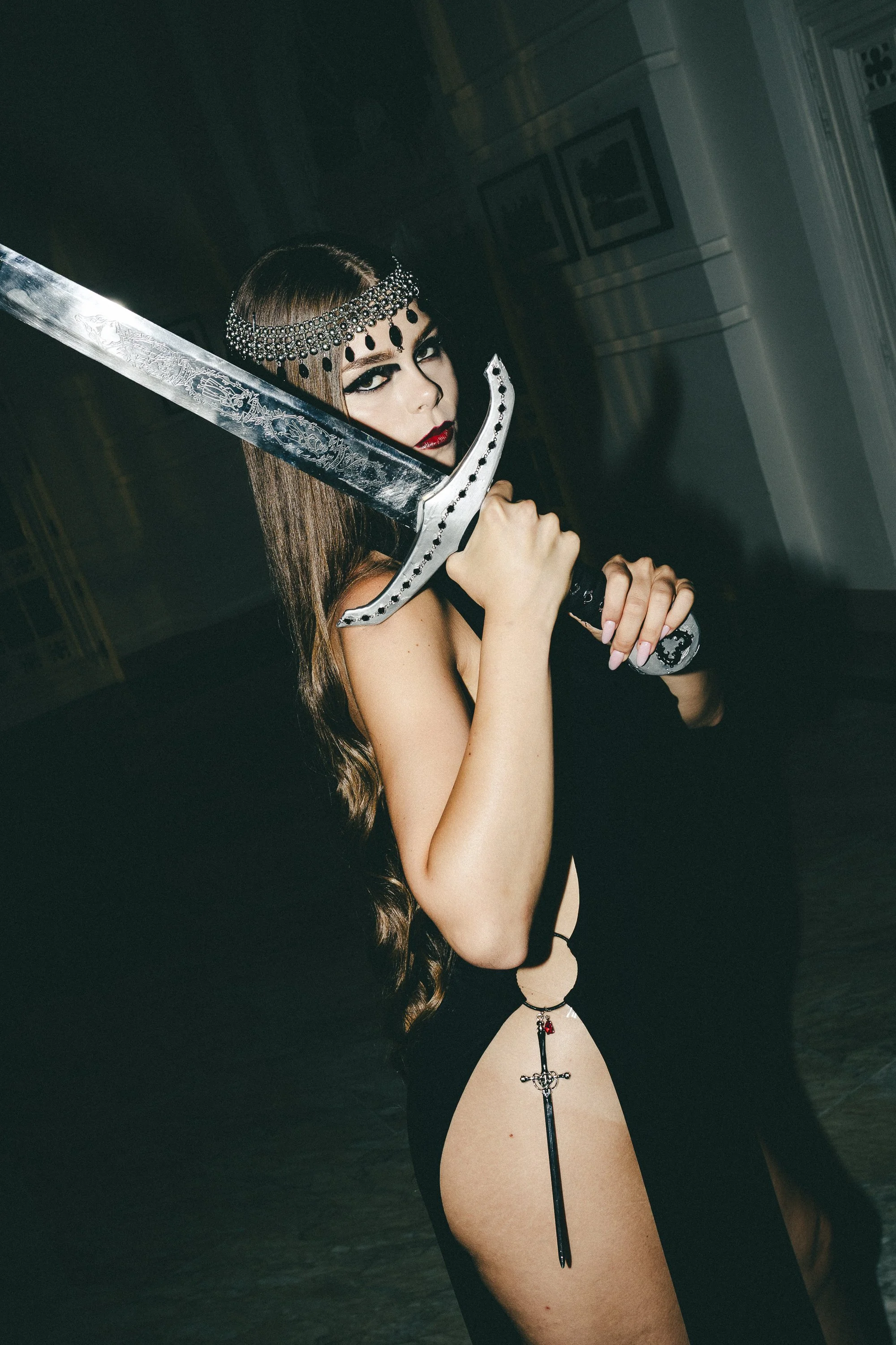 A woman with long brown hair, wearing dark makeup and a jeweled headband, holding a large sword near her face, dressed in a revealing black outfit with a thigh-high slit, in an indoor setting.