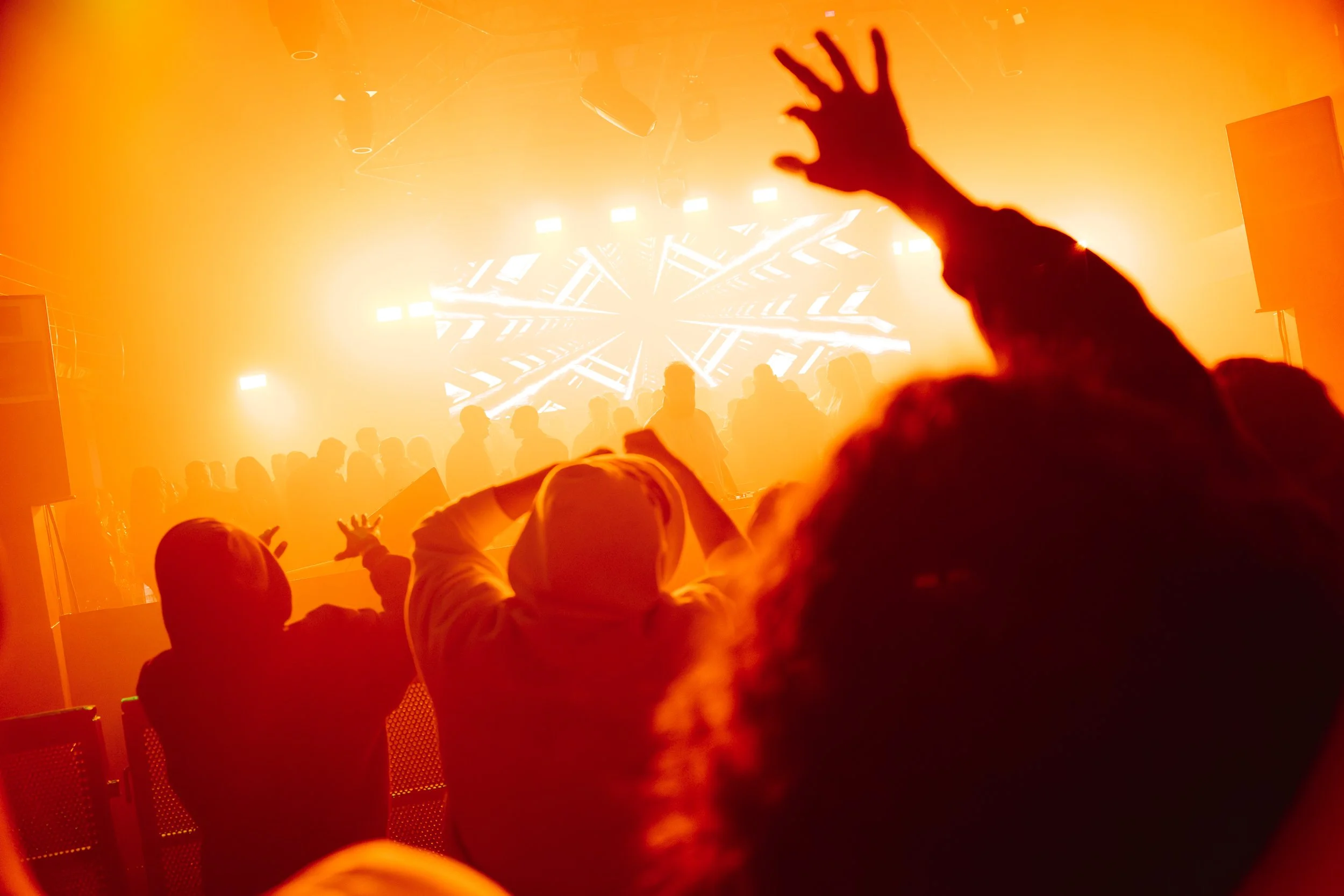 Crowd of people dancing and enjoying a concert or music event with bright orange and yellow lights and a large LED screen in the background.