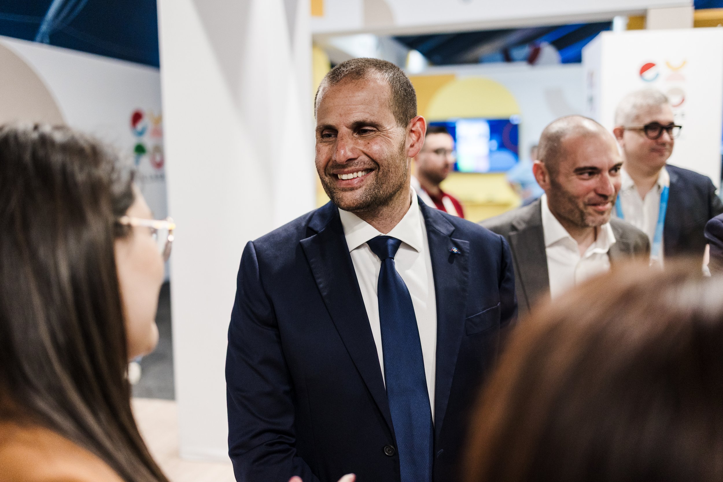 A man in a dark blue suit and tie smiling and talking to a woman with glasses at a professional event or conference, with other men and people in the background.