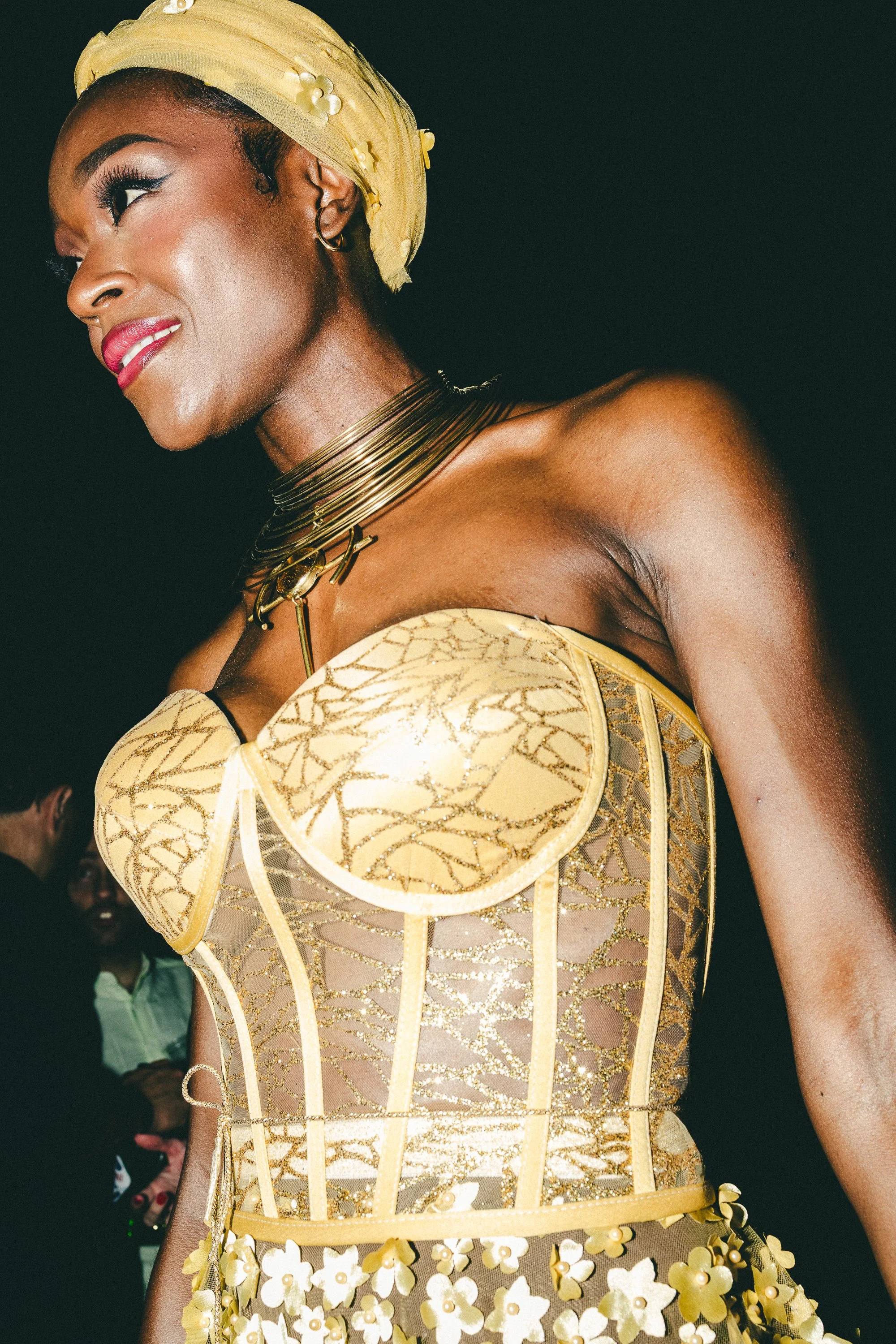 Close-up of a woman wearing a yellow and gold strapless dress with gold and floral details, adorned with multiple gold necklaces and a yellow headwrap, at a social event against a dark background.