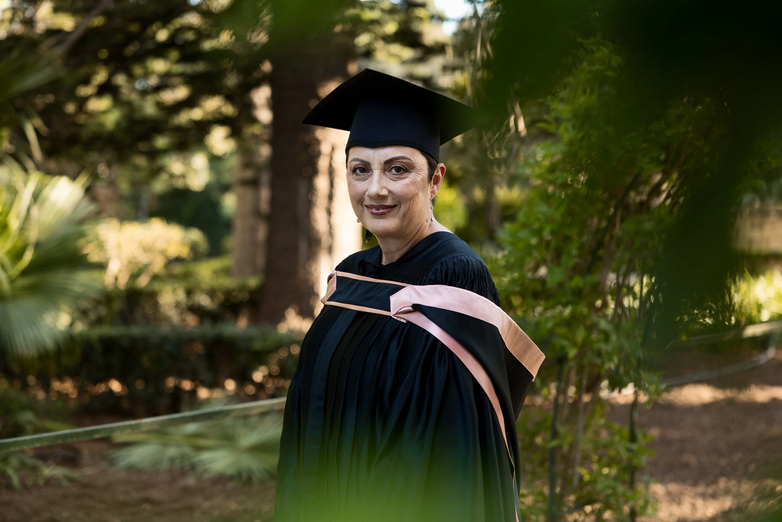 A woman in a graduation gown and cap standing outdoors among trees and greenery, smiling at the camera.