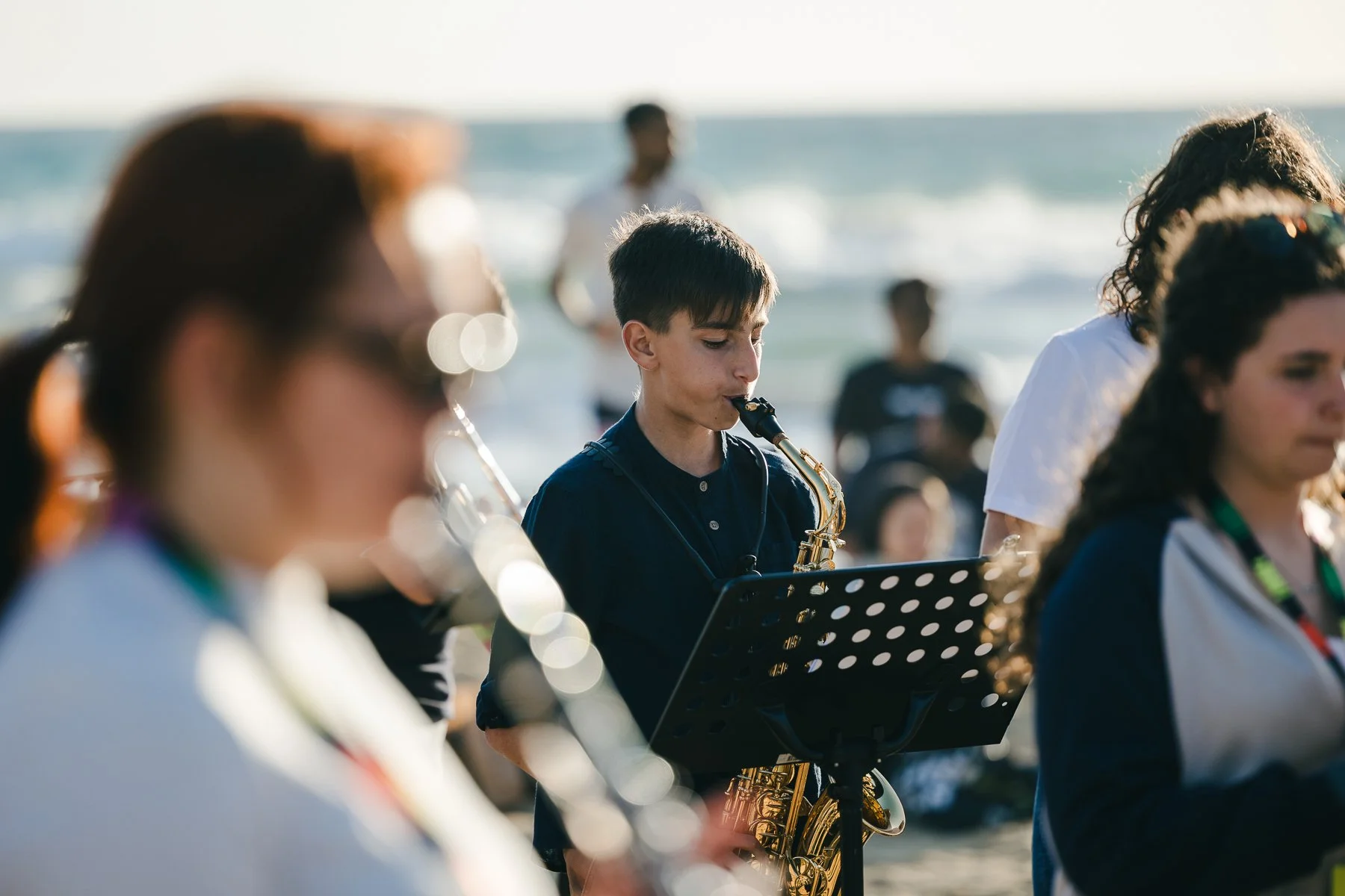 Young boy playing saxophone at outdoor beachside gathering with other musicians