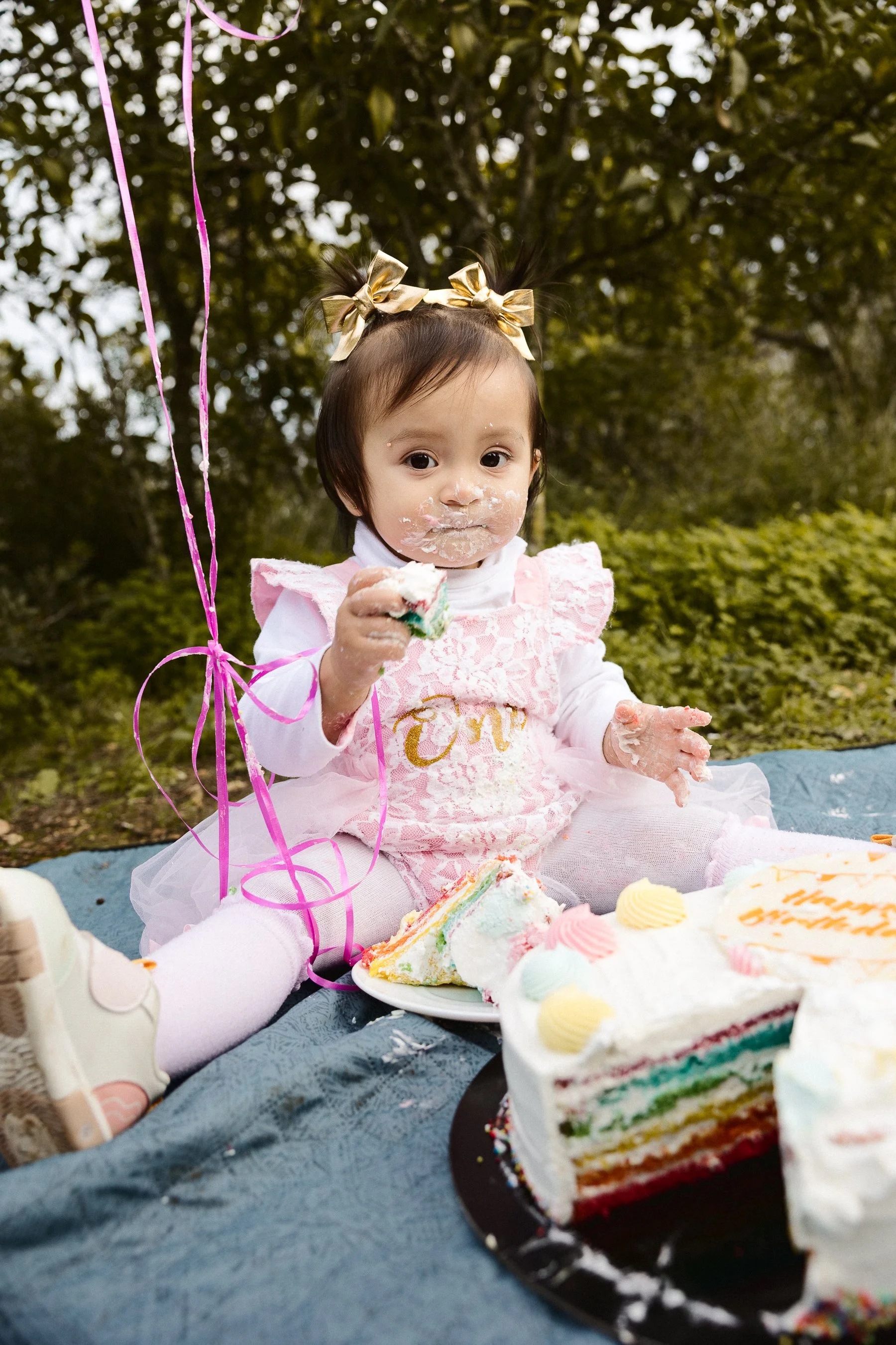 A young girl celebrating her birthday outdoors, sitting on a blanket with a rainbow cake and slice in her hand. She has cake on her face and is surrounded by balloons and snacks, with trees in the background.