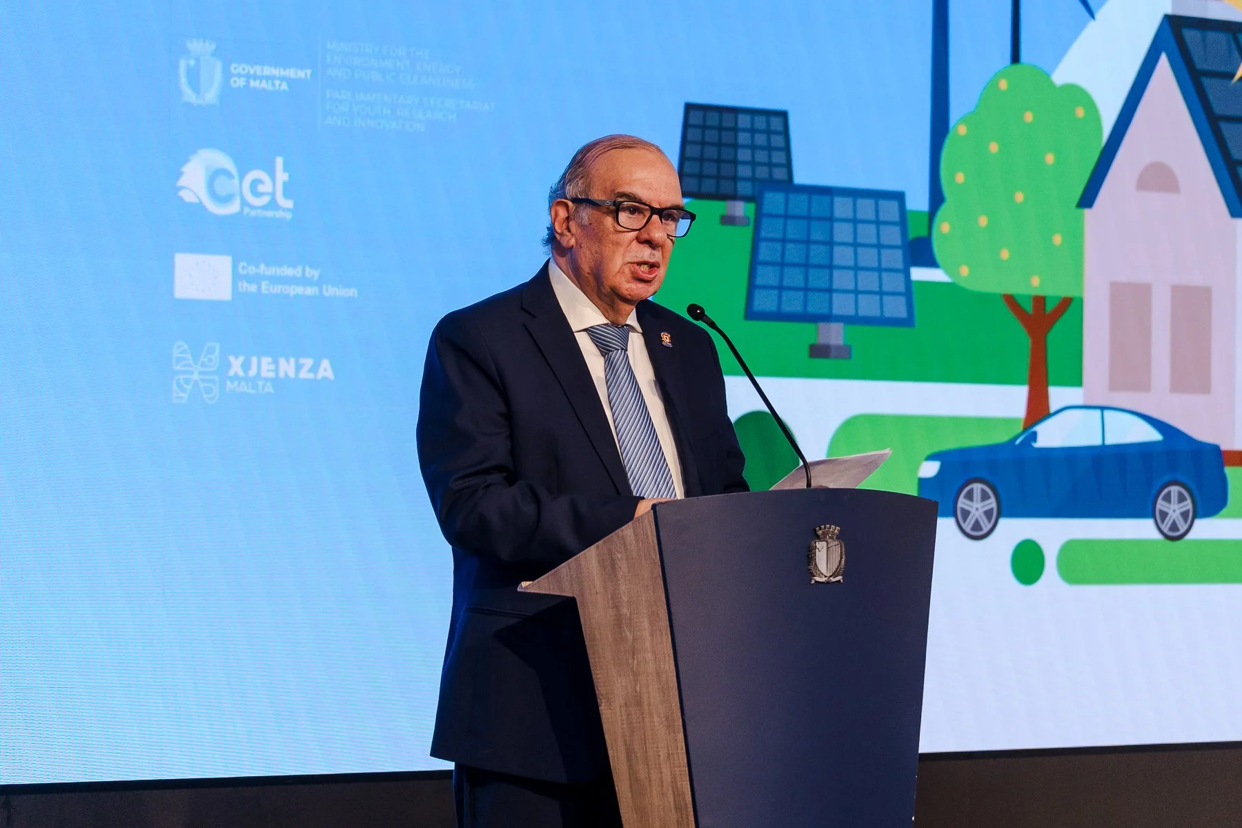 A man in a suit and tie speaking at a podium with a blue and green background depicting solar panels, trees, a house, and a blue car, with various logos and text related to Malta's government and environmental initiatives.