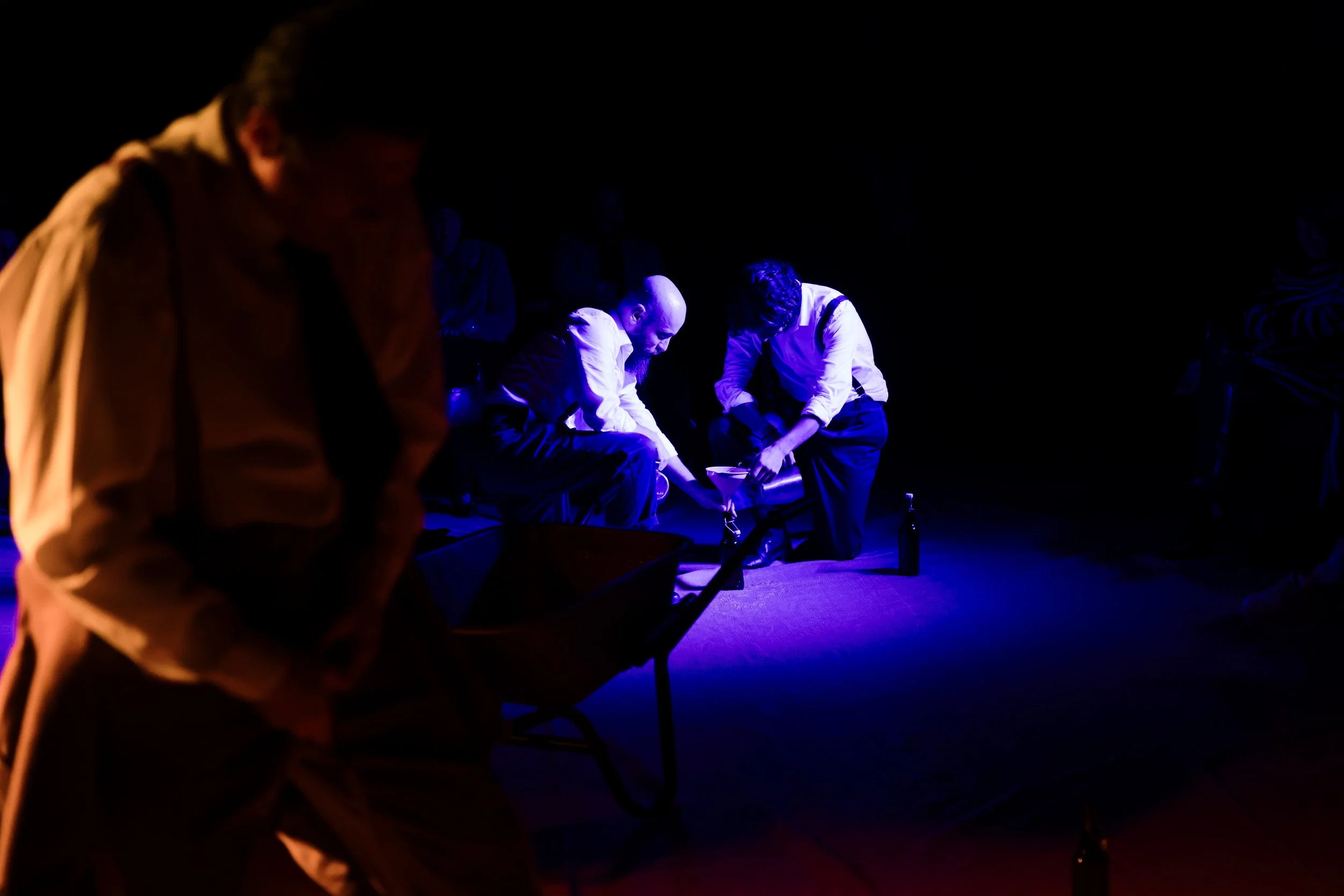 Two men in white shirts and dark pants are kneeling on the floor, illuminated by blue light, appearing to engage with a bottle and some objects. In the foreground, a blurred person in a beige shirt is walking past. Other faint figures are in the back