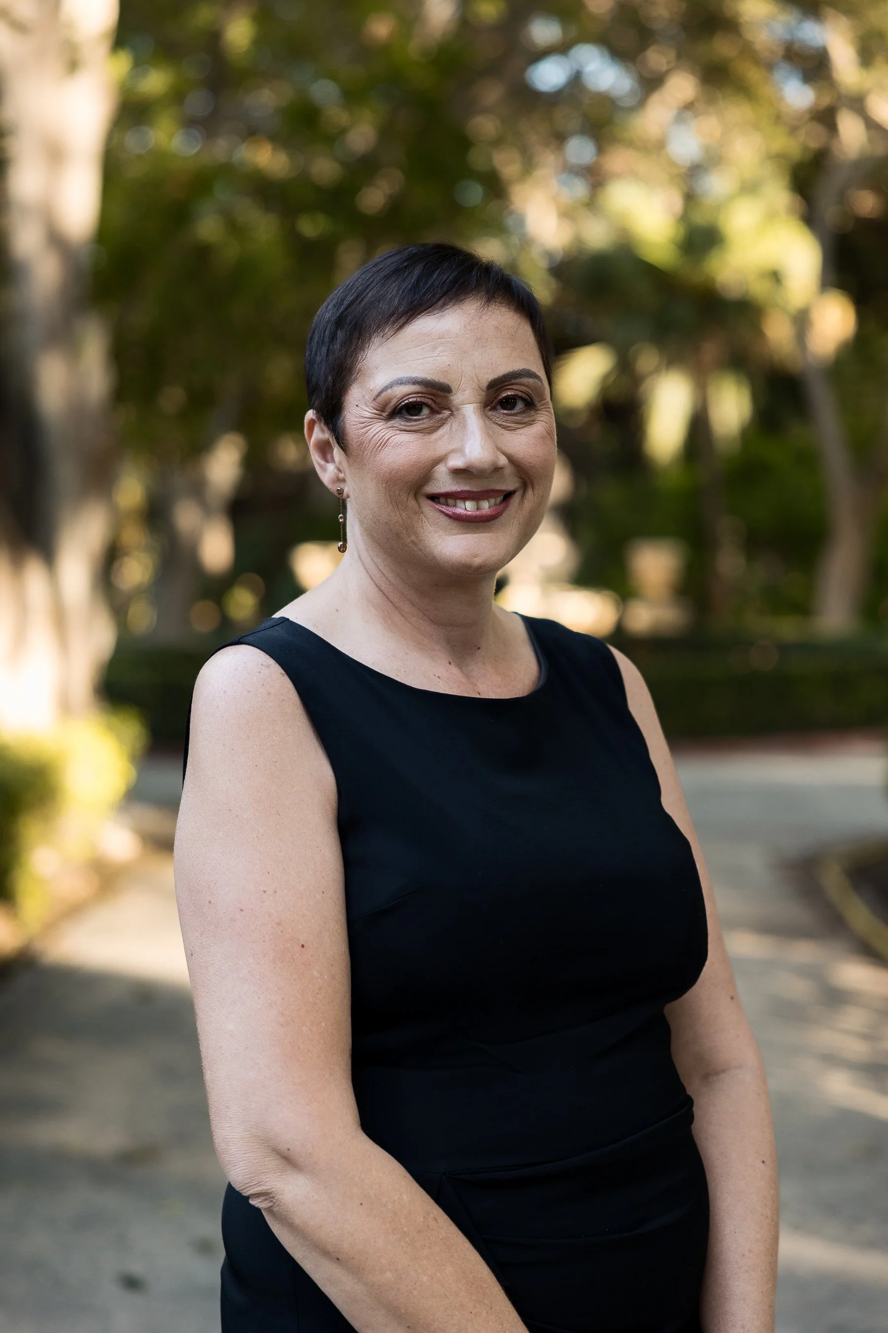 A woman with short dark hair wearing a sleeveless black dress, standing outdoors in a park-like setting with trees and greenery in the background, smiling at the camera.