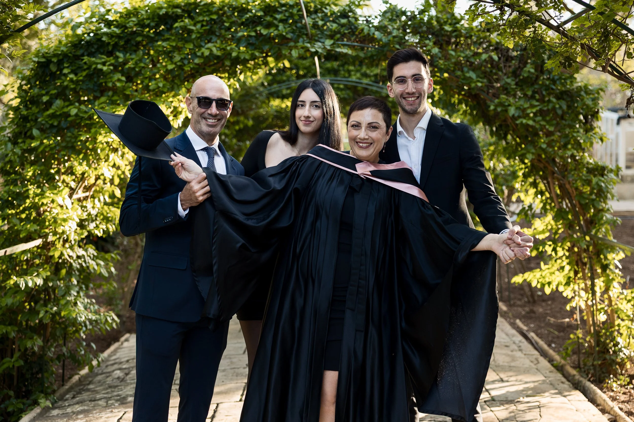 Group of four people, including a woman in graduation gown, celebrating outdoors on a pathway surrounded by greenery, with one person holding a graduation cap.