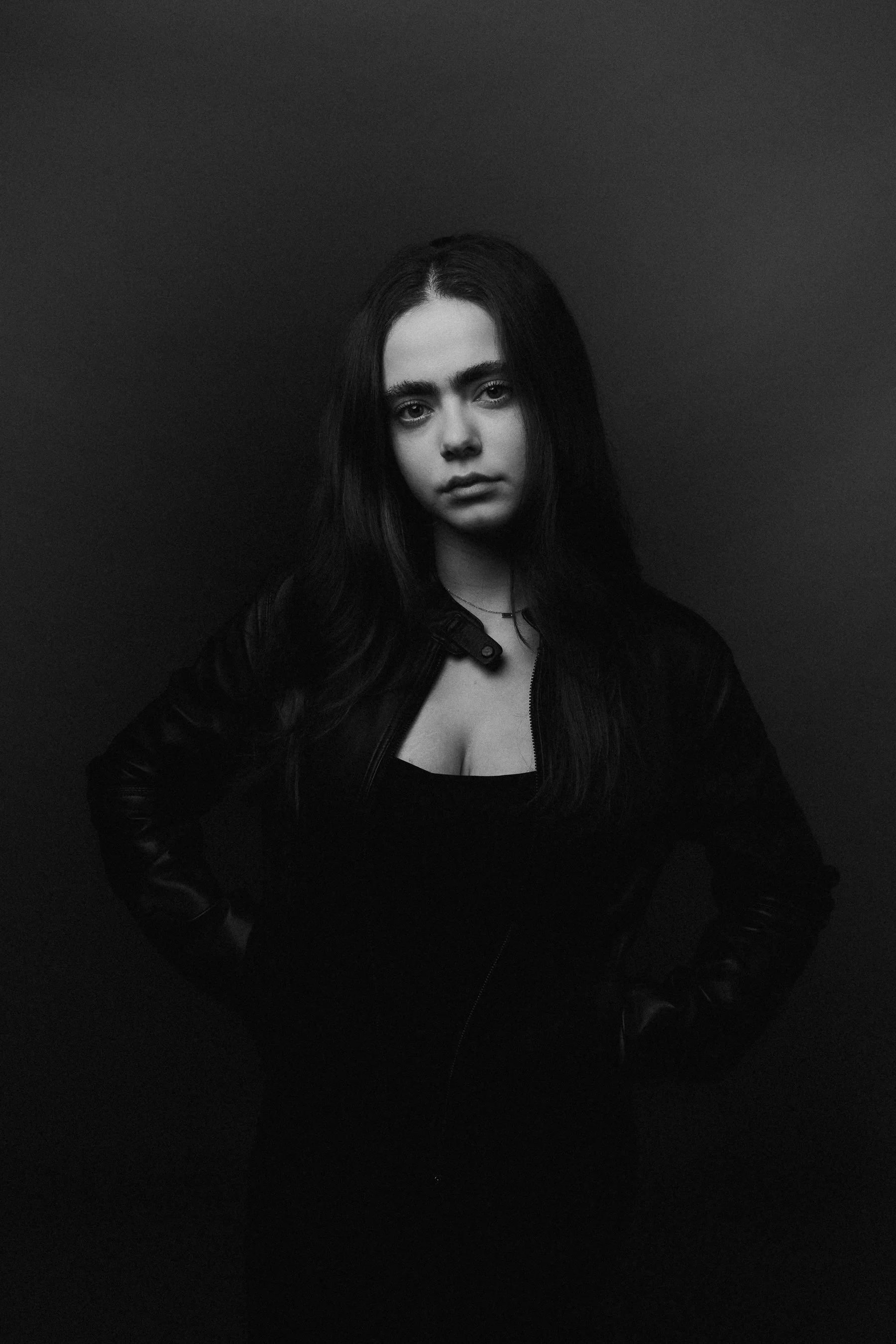 Black and white photo of a young woman with long dark hair, wearing a leather jacket and a black top, standing against a plain dark background with her hands on her hips.