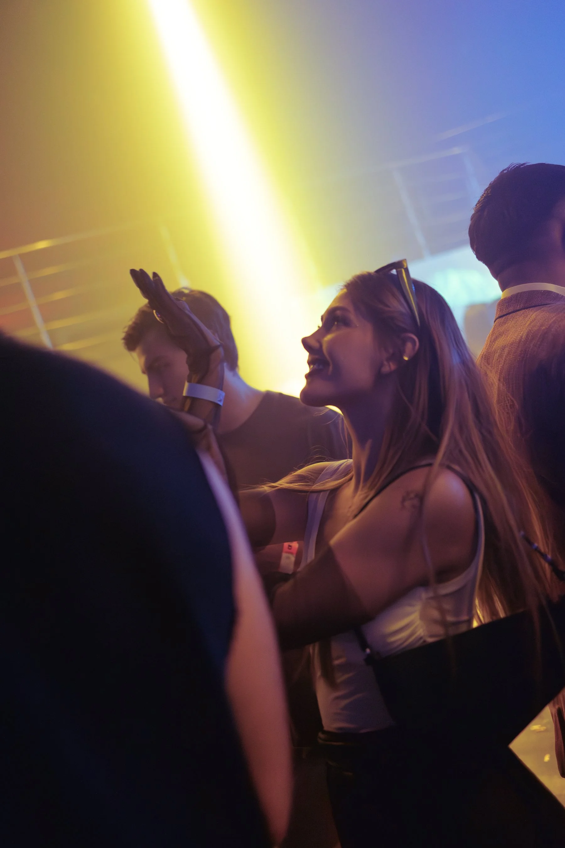 A woman with long hair and sunglasses on her head smiling and talking at a crowded party or club, with multiple people around her and colorful lighting.