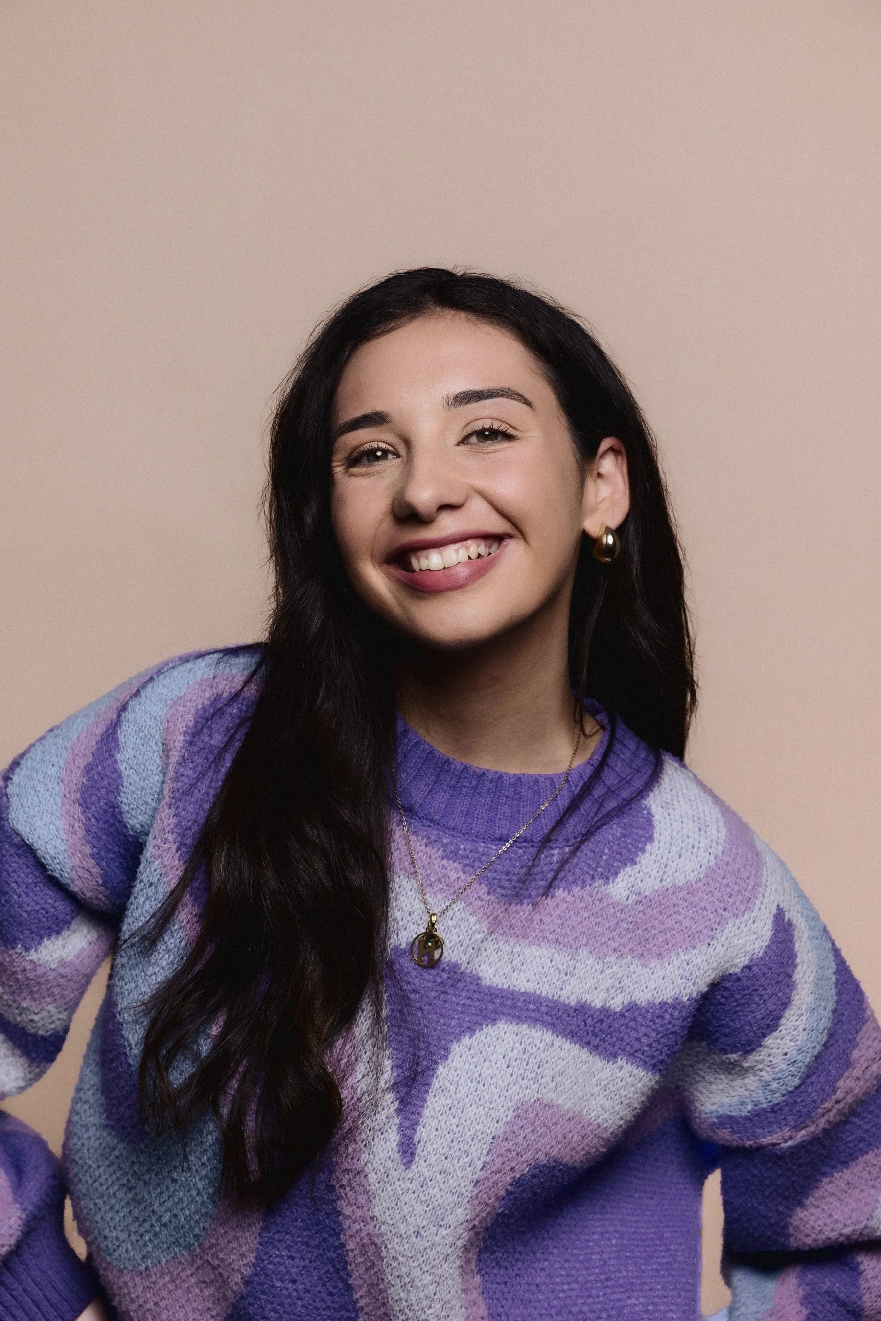 A young woman with long dark hair, smiling with teeth showing, wearing a purple and lavender patterned sweater and gold jewelry against a plain beige background.