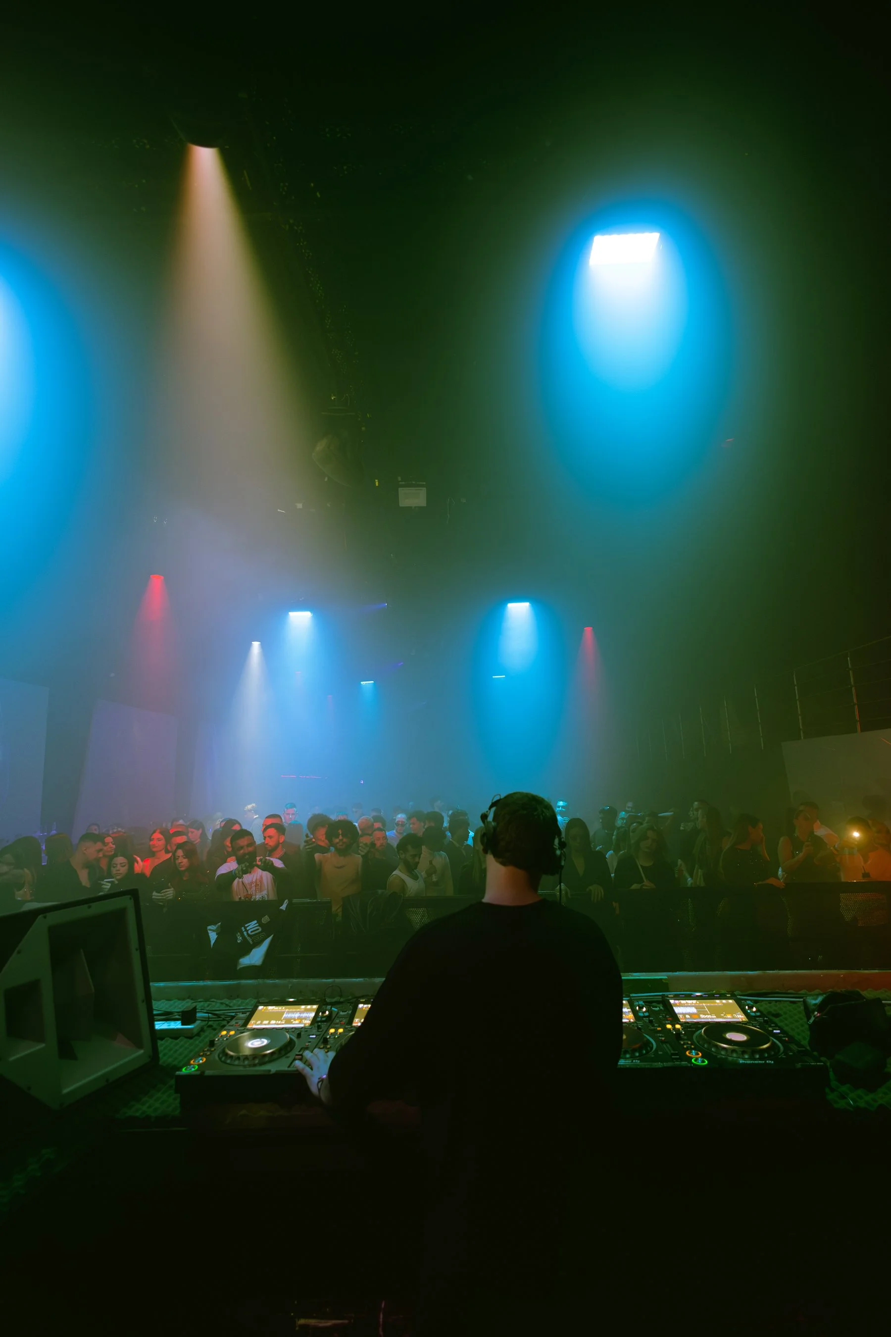 A DJ performs at a nightclub with colorful lighting, facing a crowd of people dancing and enjoying the music.