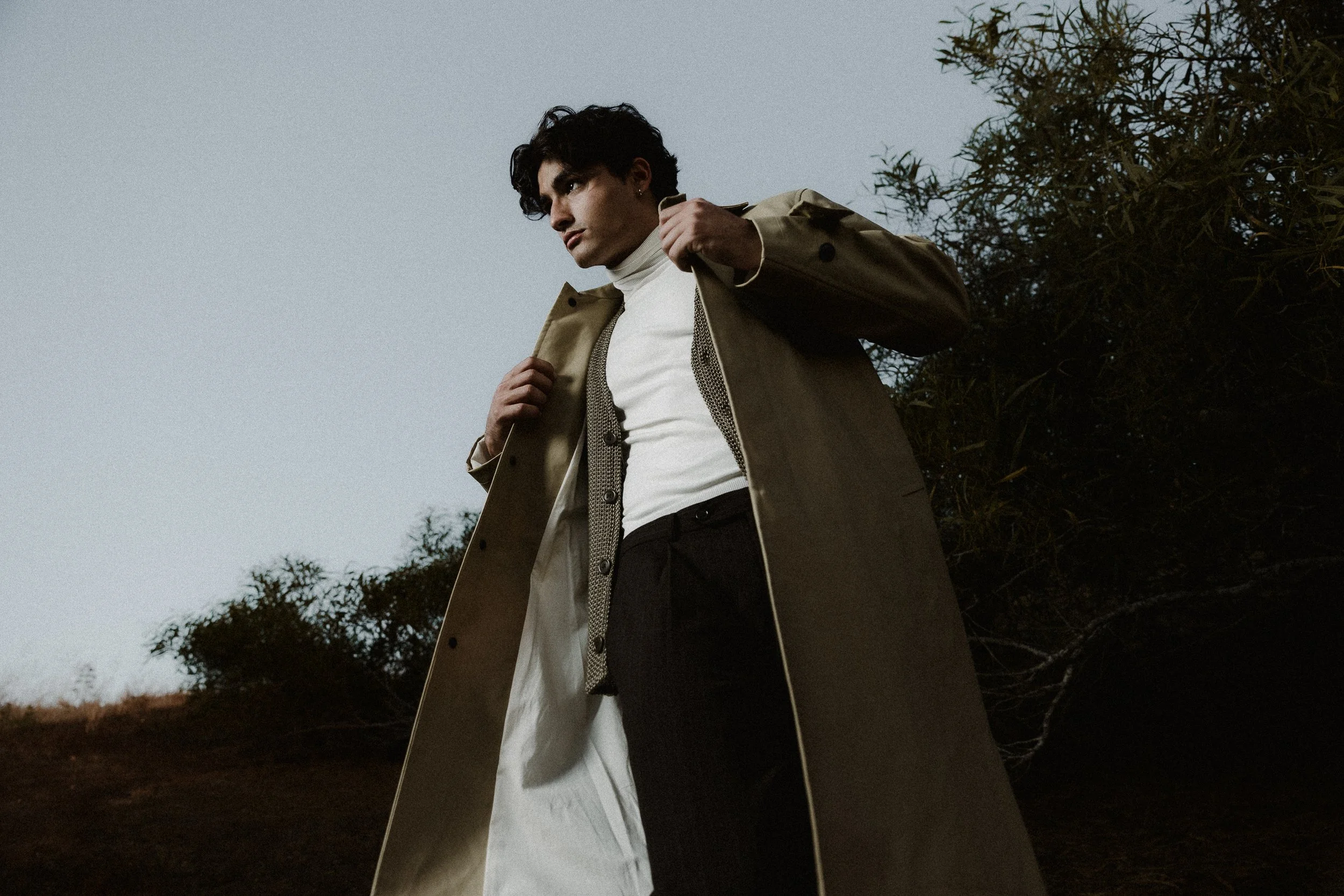 A young man with dark, curly hair, wearing a beige trench coat and white turtleneck, standing outdoors near trees under an overcast sky.
