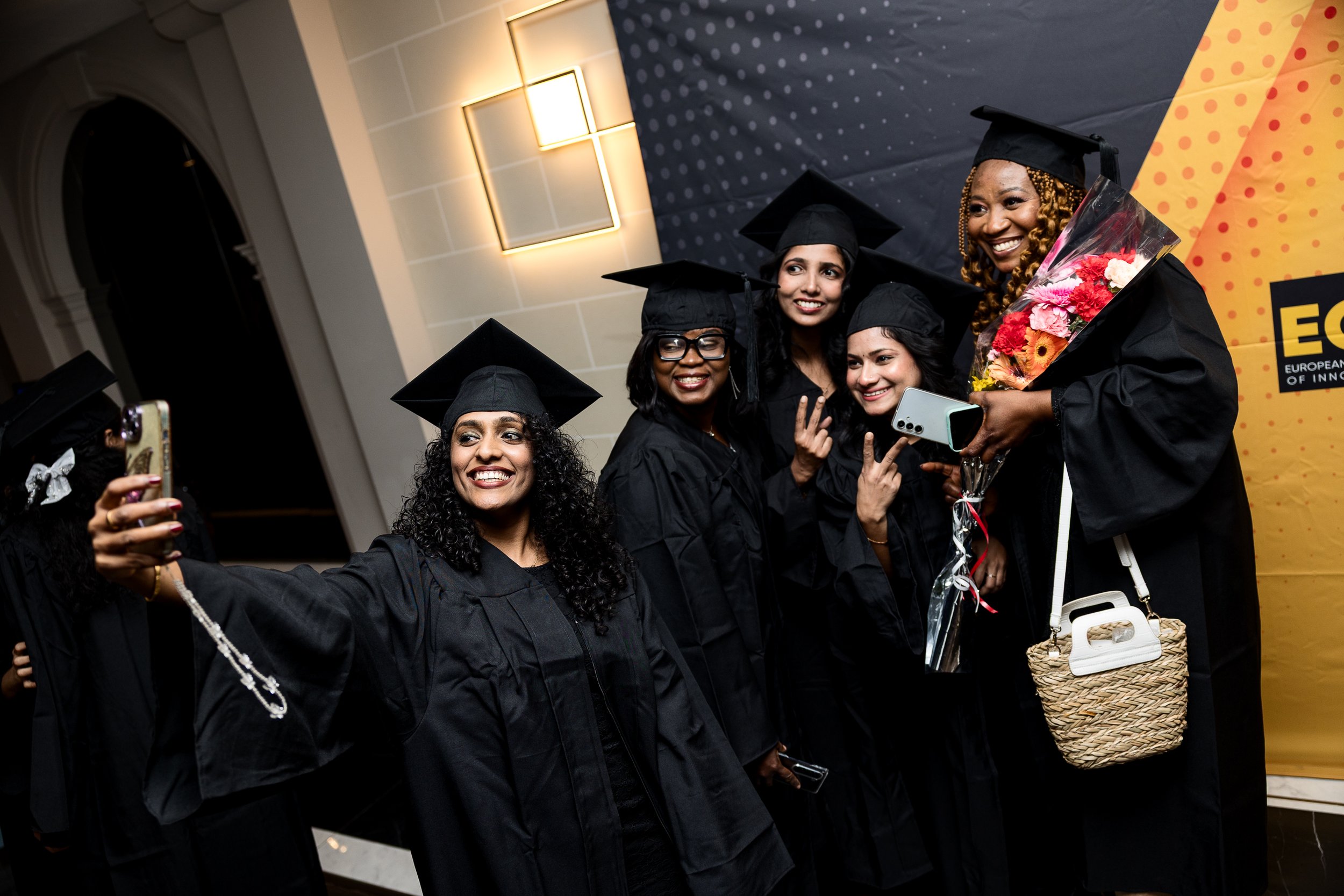 Group of women wearing black graduation gowns and caps taking a selfie at a graduation ceremony.