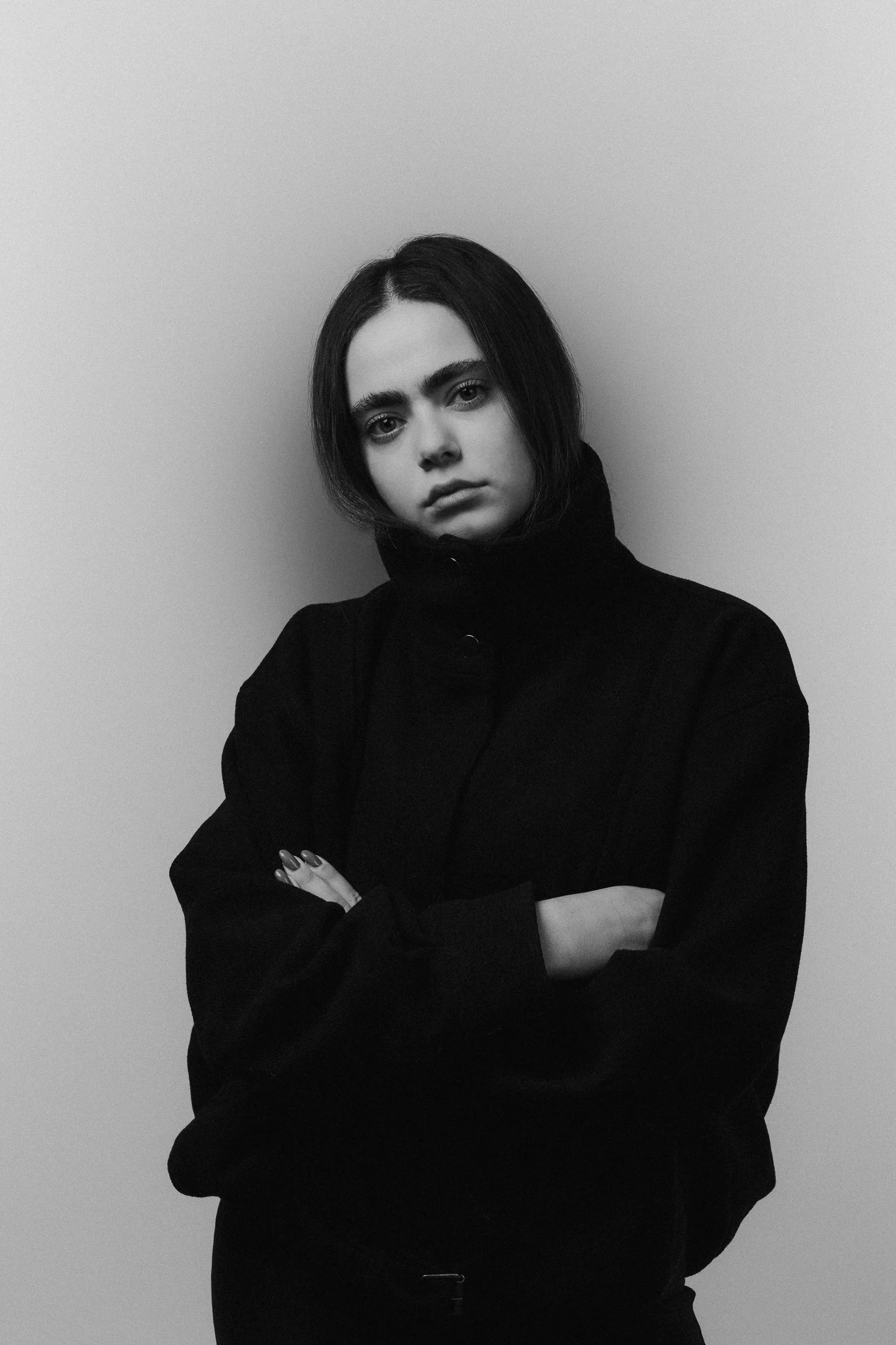 A young woman with short dark hair and arched eyebrows, wearing a black high-collared jacket, standing with arms crossed against a plain light-colored wall.