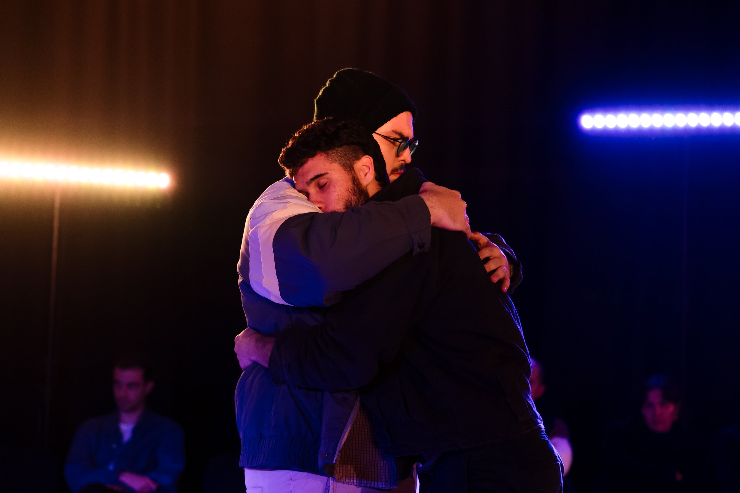 Two young men hugging each other in an emotional moment at night, illuminated by colorful lights.