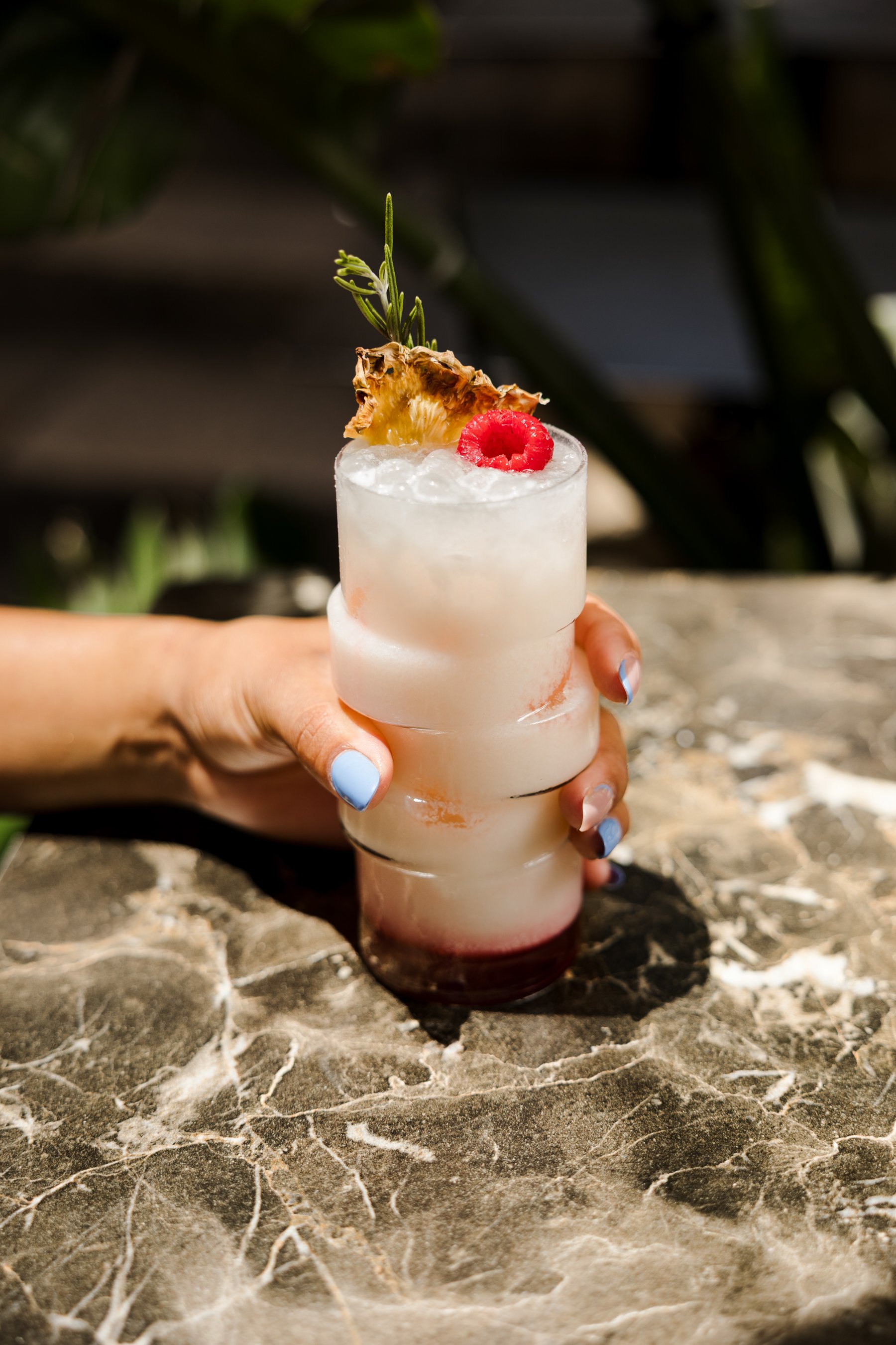A cocktail drink in a tall glass with ice, garnished with a slice of pineapple, a raspberry, and a sprig of rosemary, held by a person with blue manicure nails, on a marble surface.