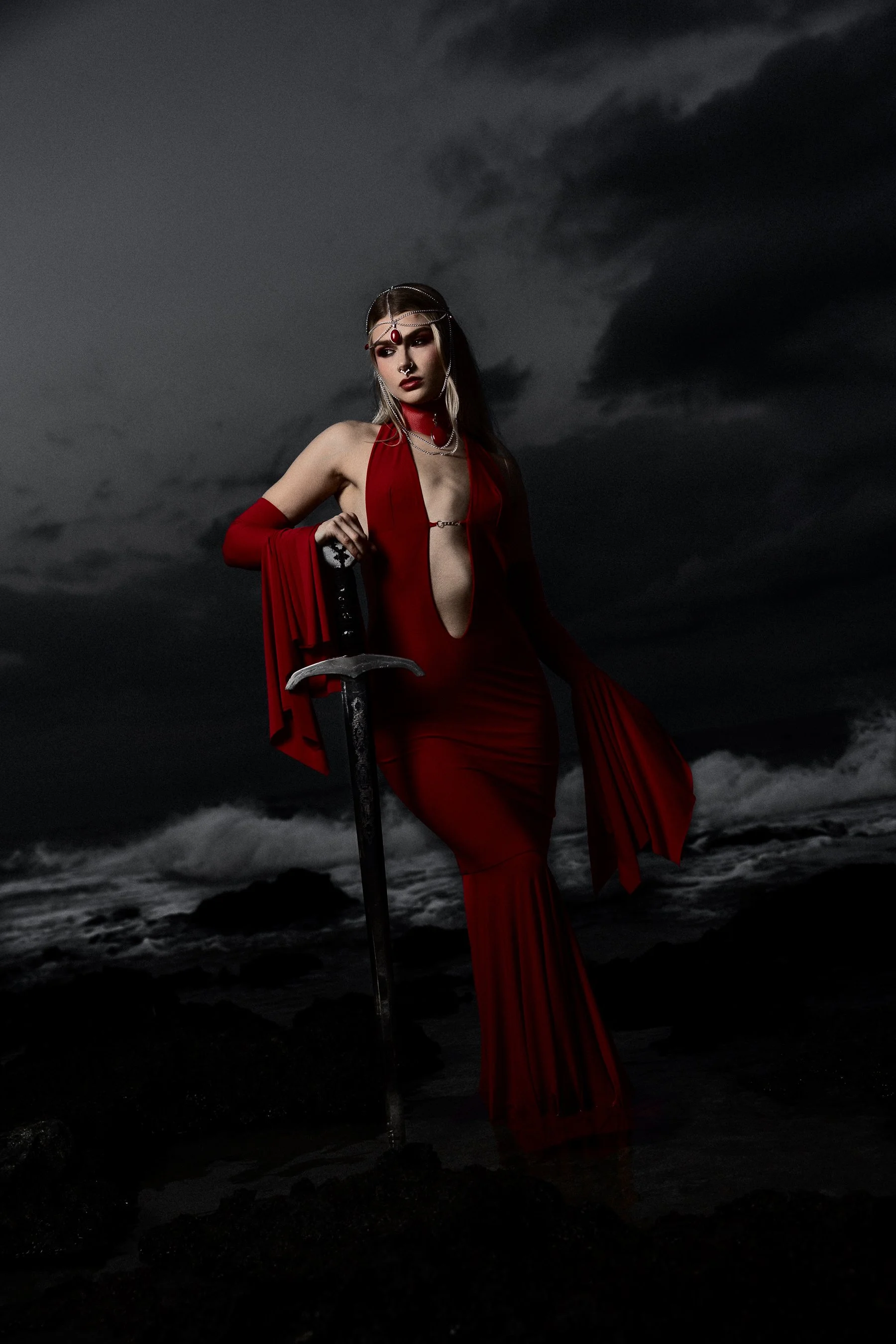 A woman in a dramatic red dress with a high slit standing on rocks by the ocean at night, holding a sword, under a dark cloudy sky.