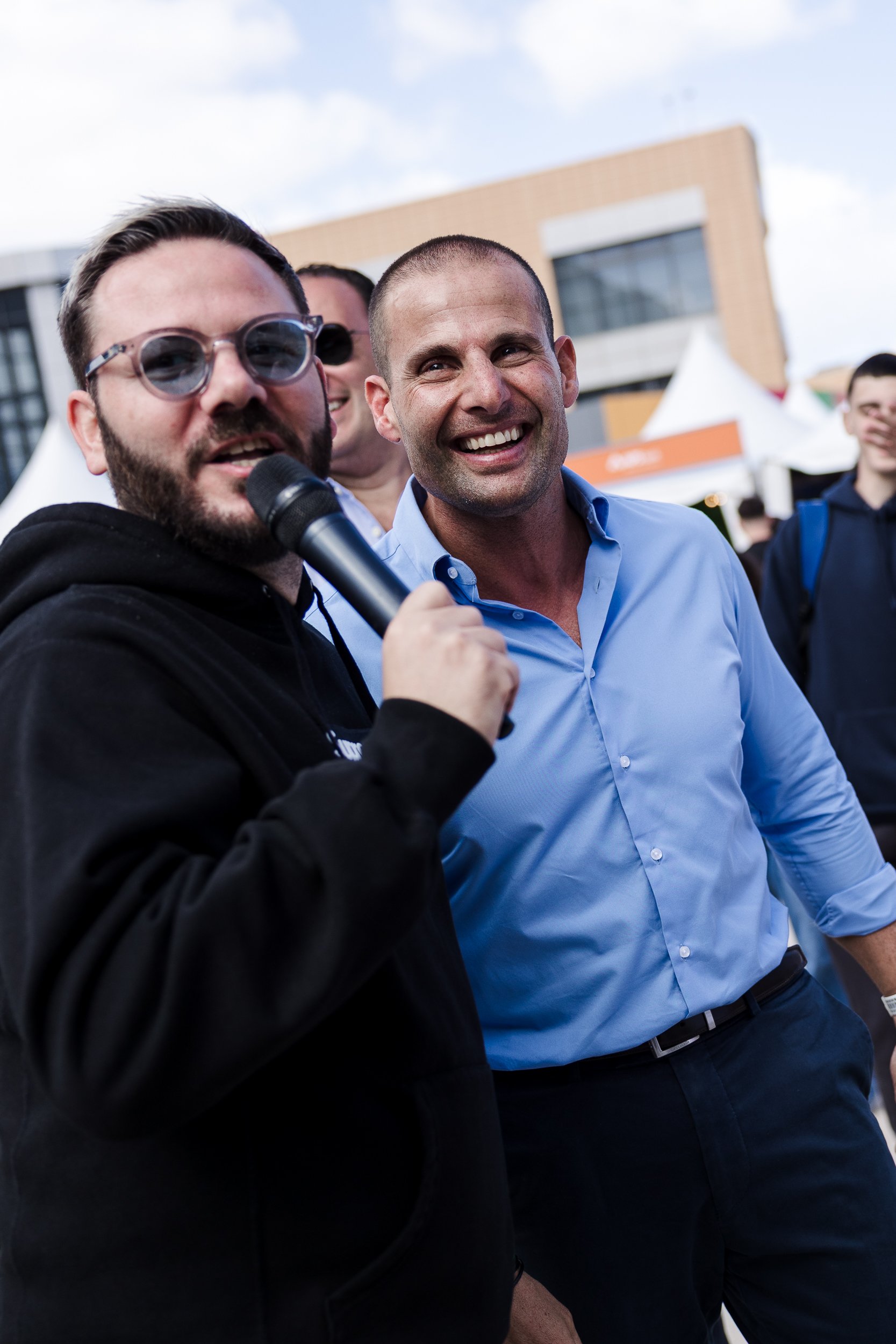 A man in a black hoodie holding a microphone, smiling, while talking at an outdoor event, with a man in a light blue shirt standing next to him.