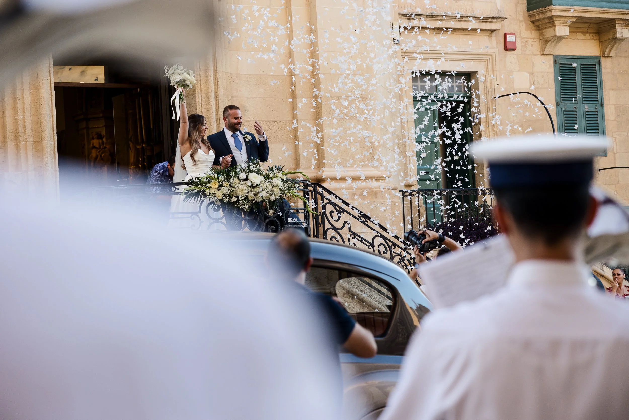 A bride and groom celebrating on a church balcony, throwing flower petals and surrounded by guests and photographers during a wedding ceremony.