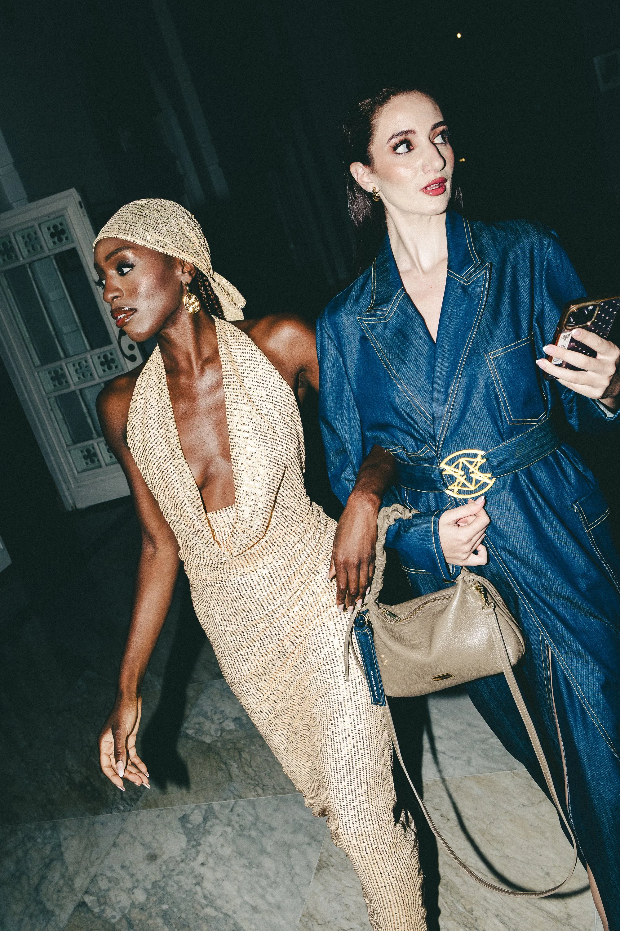 Two stylish women in fashionable outfits, one in a beige dress and headscarf, and the other in a blue denim dress with a gold belt, holding a beige handbag and a phone, walking together indoors.