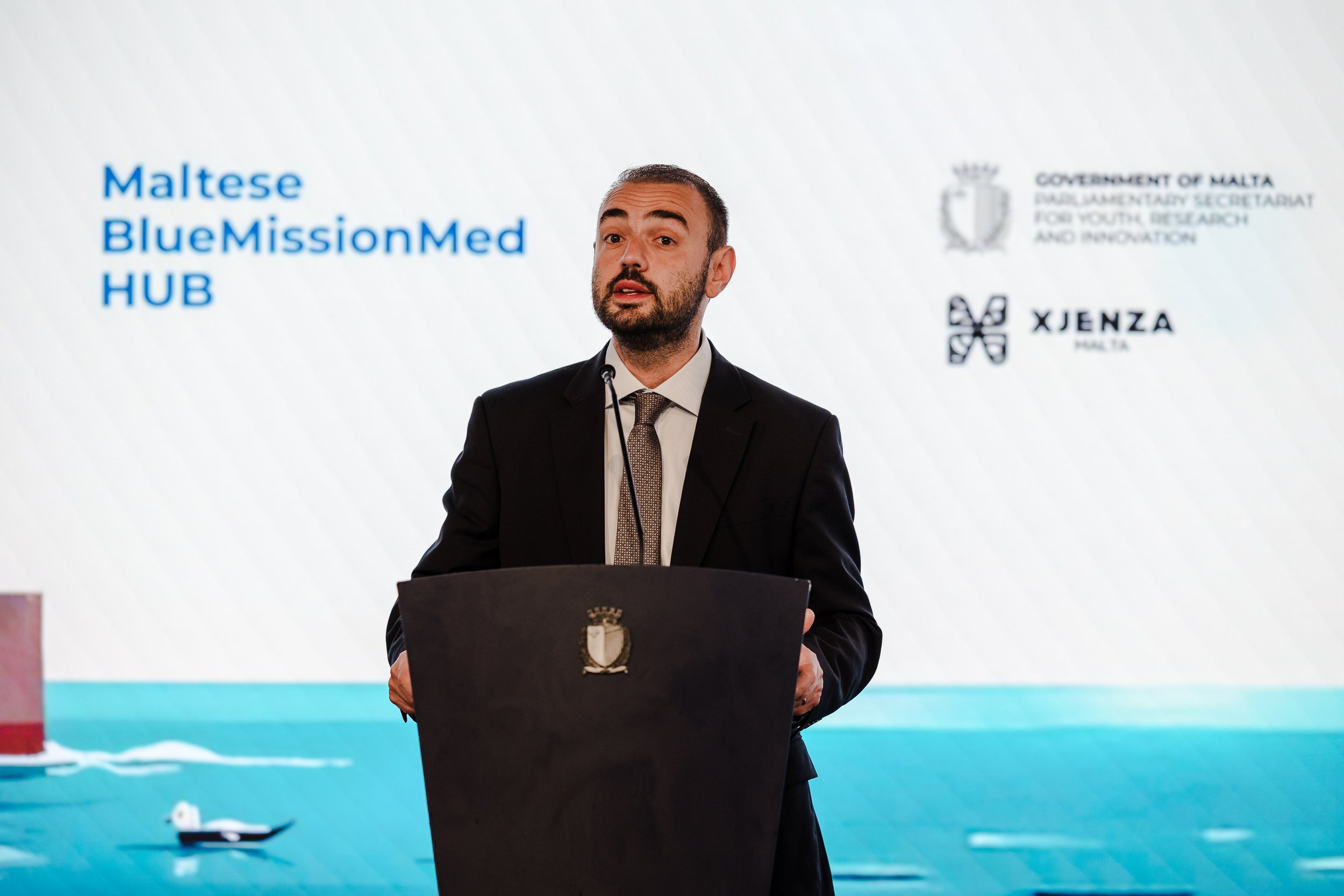 A man in a black suit and tie standing at a podium giving a speech, with a presentation slide behind him that reads 'Maltese Blue Mission Med HUB' and includes logos of the Government of Malta and other entities.