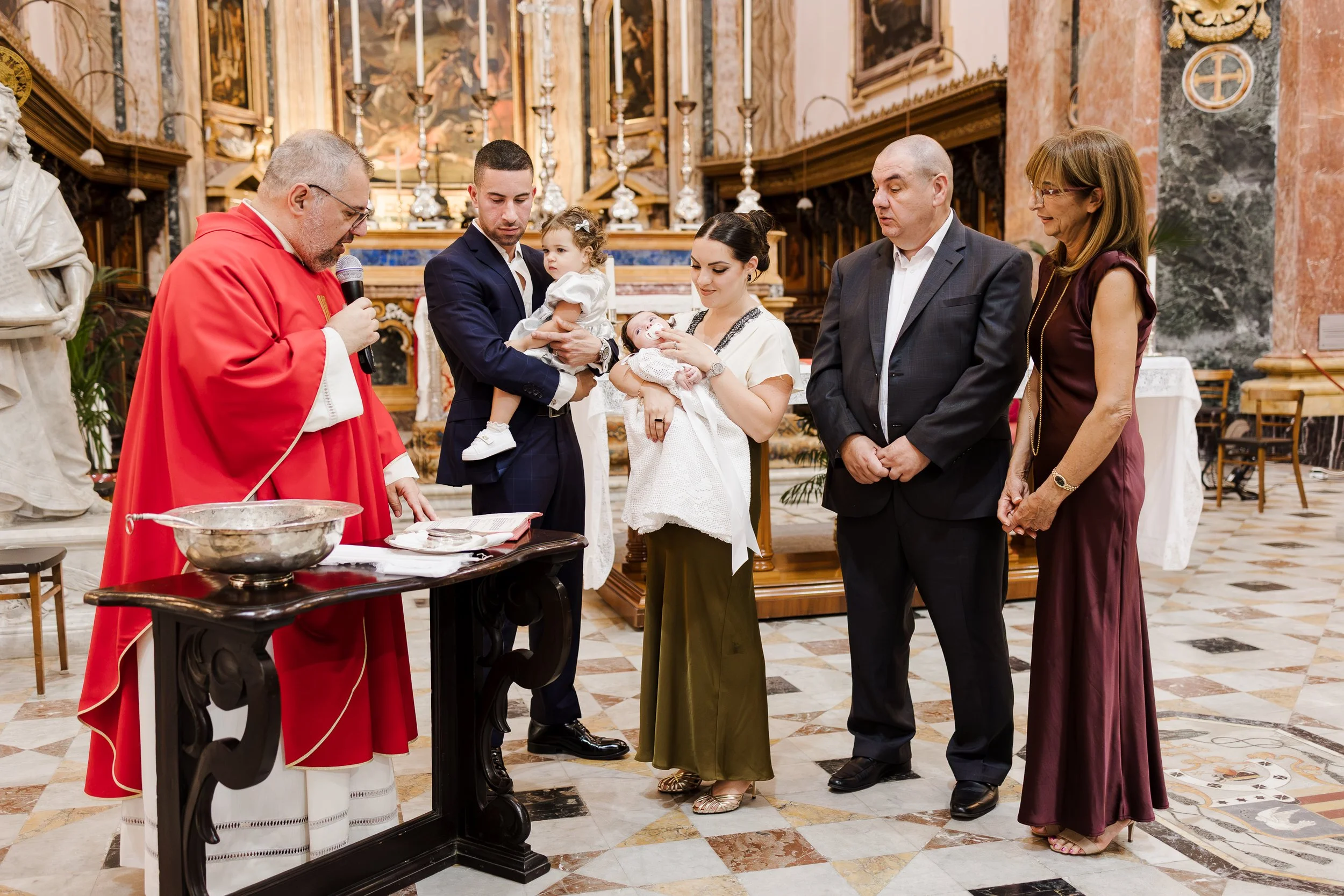 A baptism ceremony taking place inside a church with a priest, a family of six including a priest, a man, a woman, two children, and a woman, participating in the religious ritual.