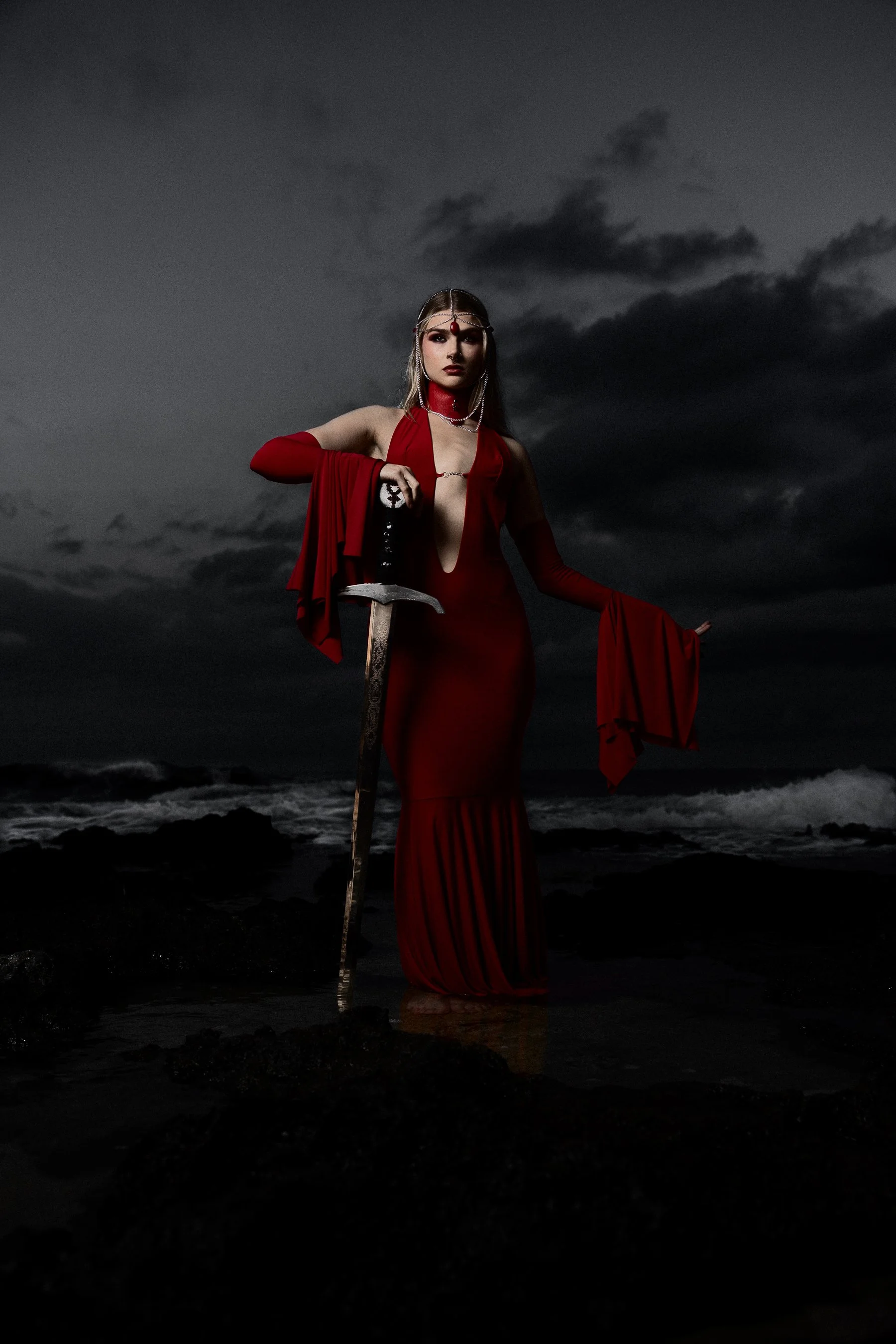 A woman dressed in a flowing red gown standing in shallow water on a rocky beach at dusk or night, holding a sword, with dark clouds in the sky behind her.