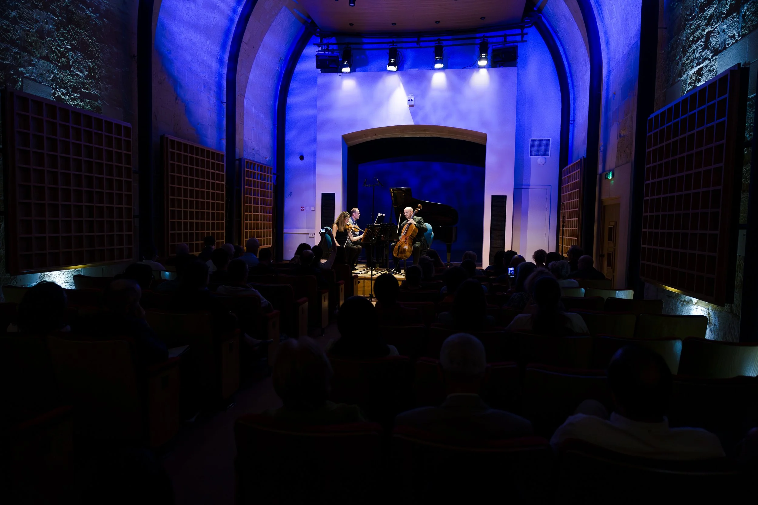 A live classical music concert with three musicians performing on stage, illuminated by blue and white lighting, in front of an audience seated in a dimly lit auditorium.