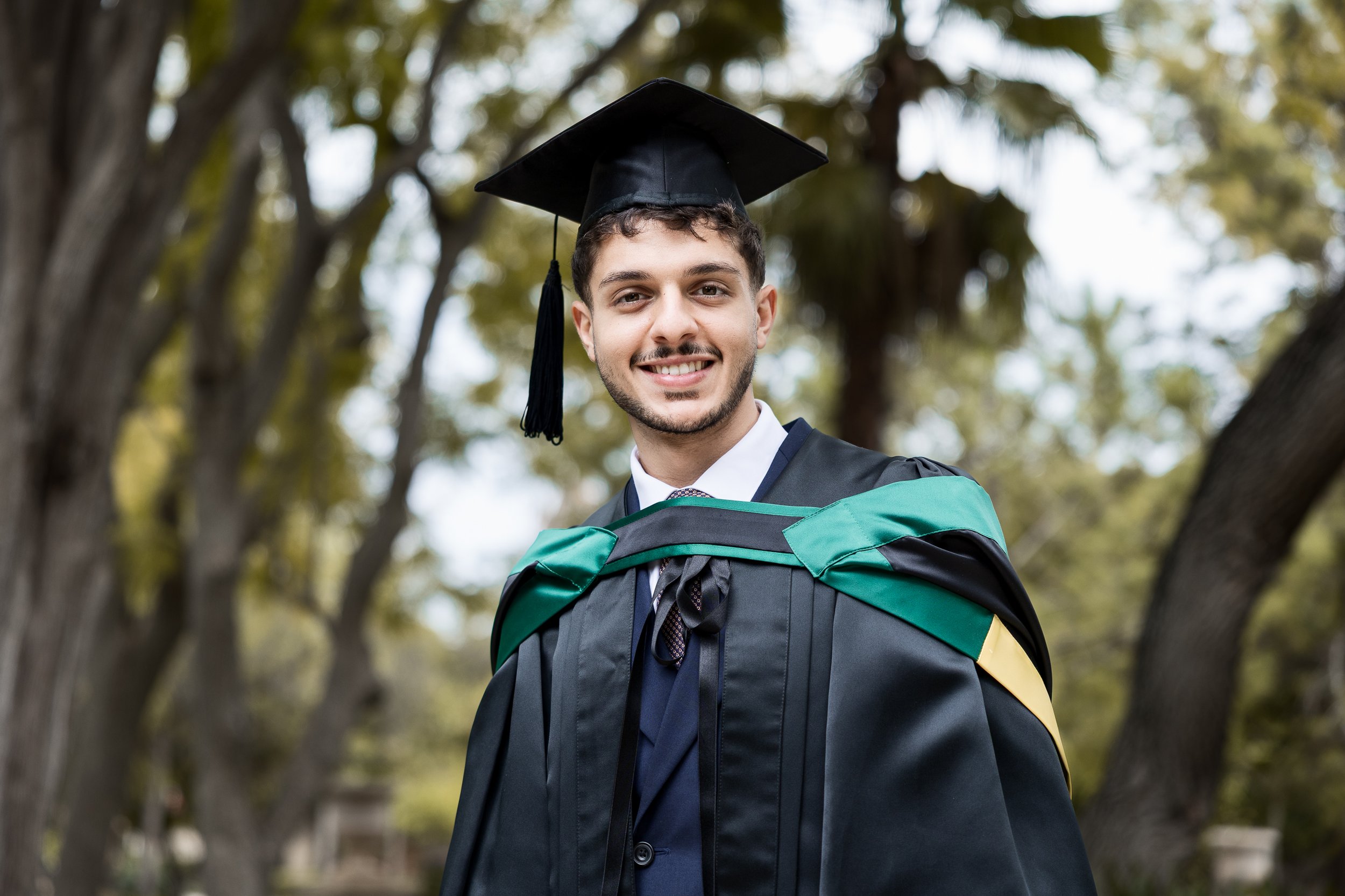 Young man in graduation cap and gown smiling outdoors with trees in background.