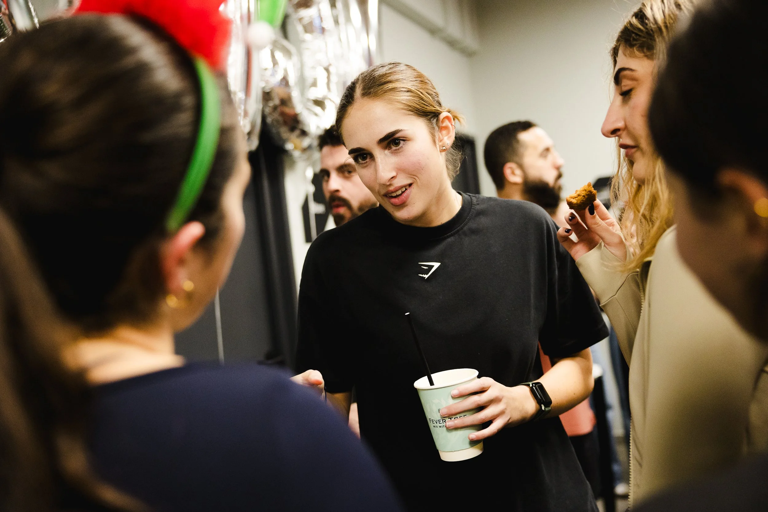 A group of people socializing indoors, with a woman at the center holding a cup and engaged in conversation.