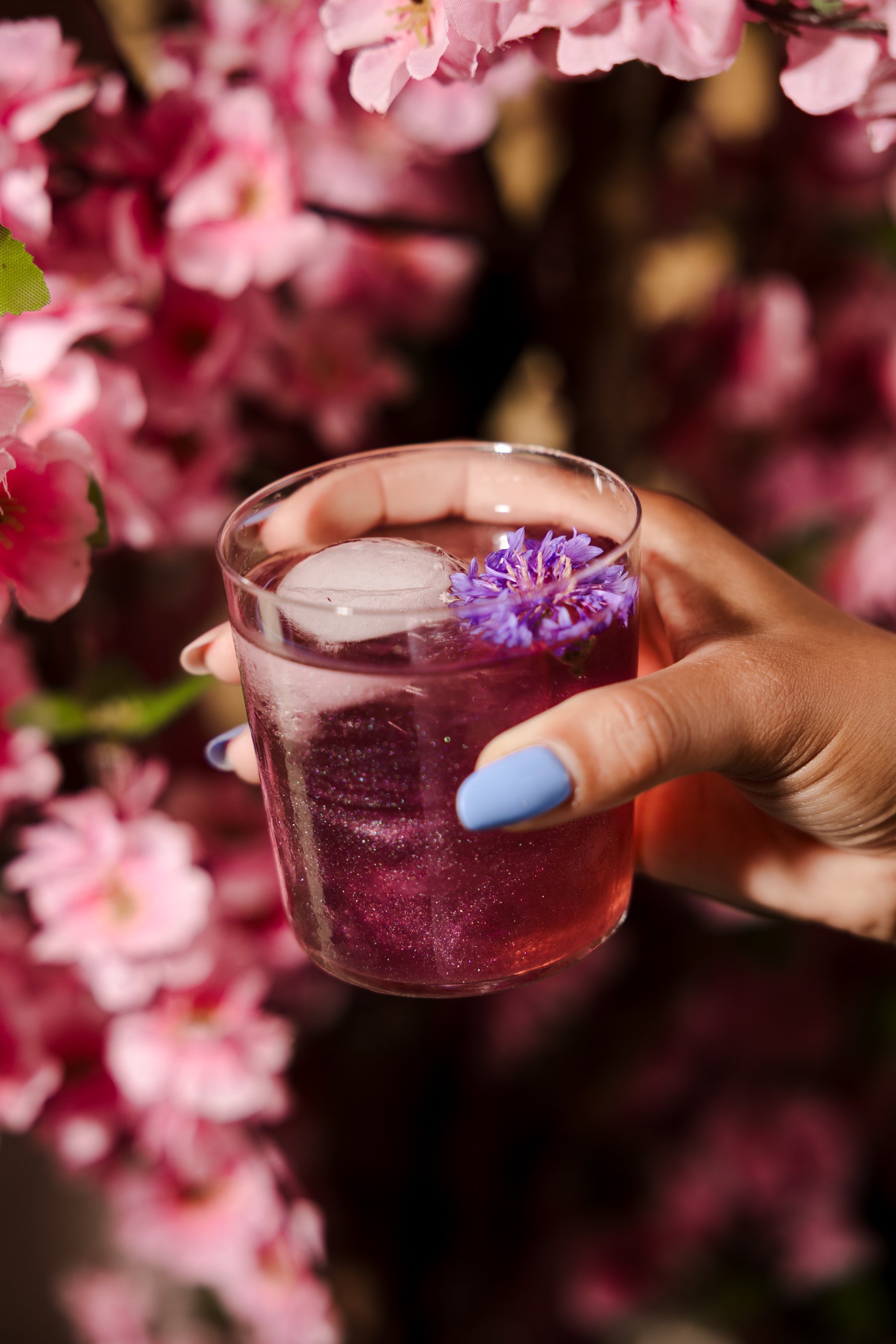 A hand with blue-painted nails holding a glass of pink drink with a purple flower floating on top, background of pink blossoms.