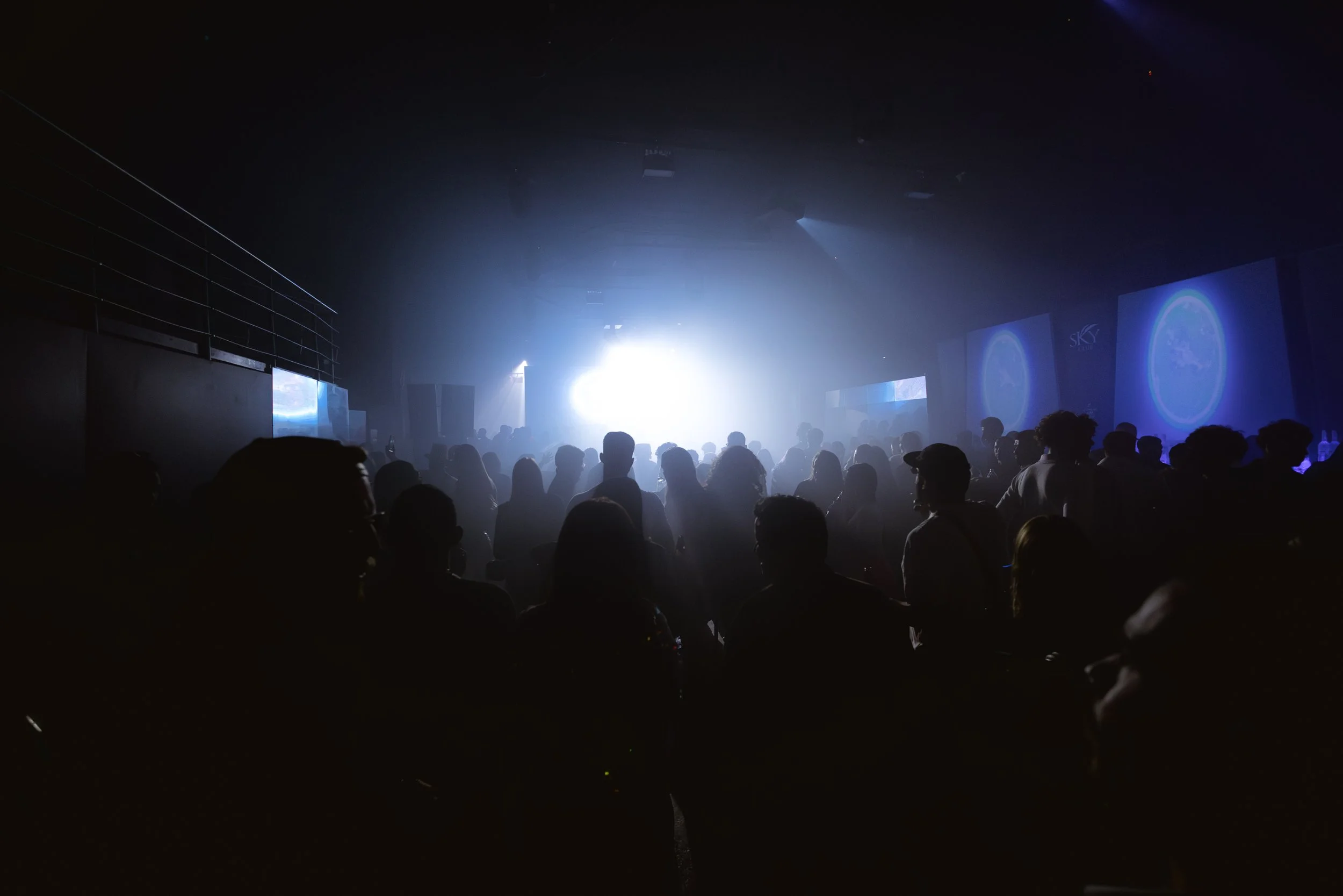 People dancing and socializing in a dark nightclub with bright stage lights and large screens displaying graphics.