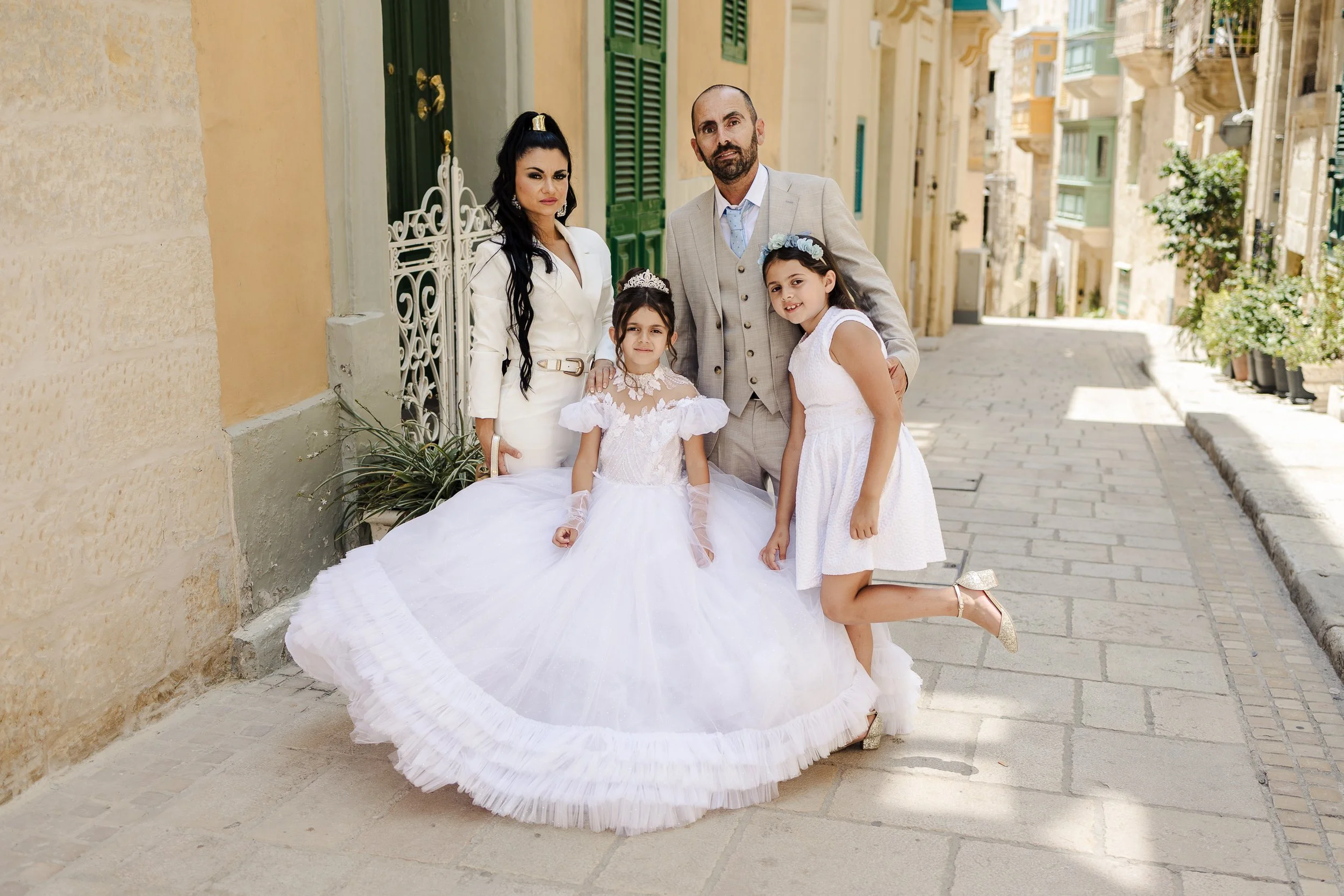 Family of four dressed in formal attire on a cobblestone street with colorful European-style buildings.