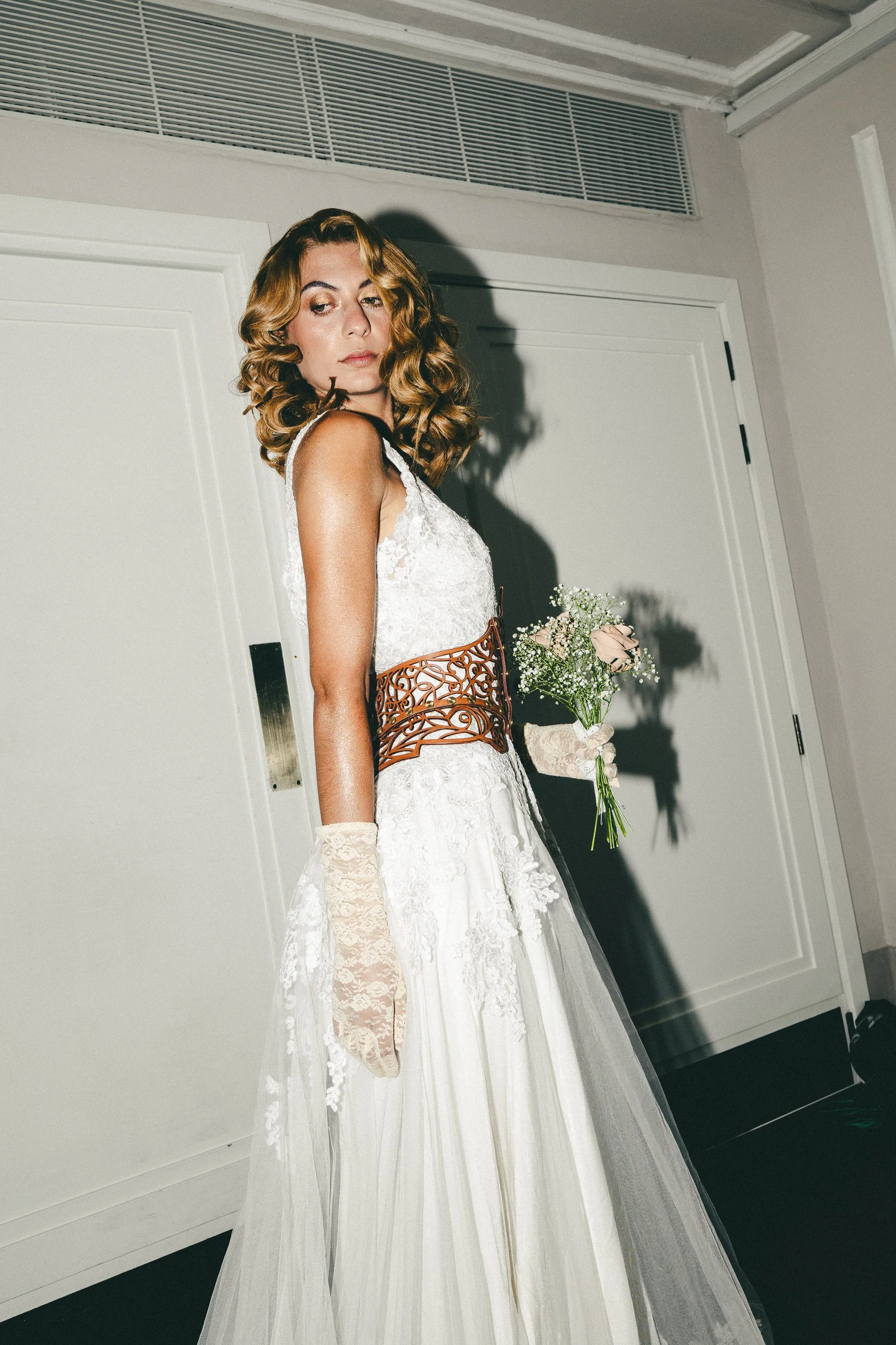 Woman in lace wedding dress holding a bouquet of flowers, standing against a plain white wall with a shadow cast behind her.