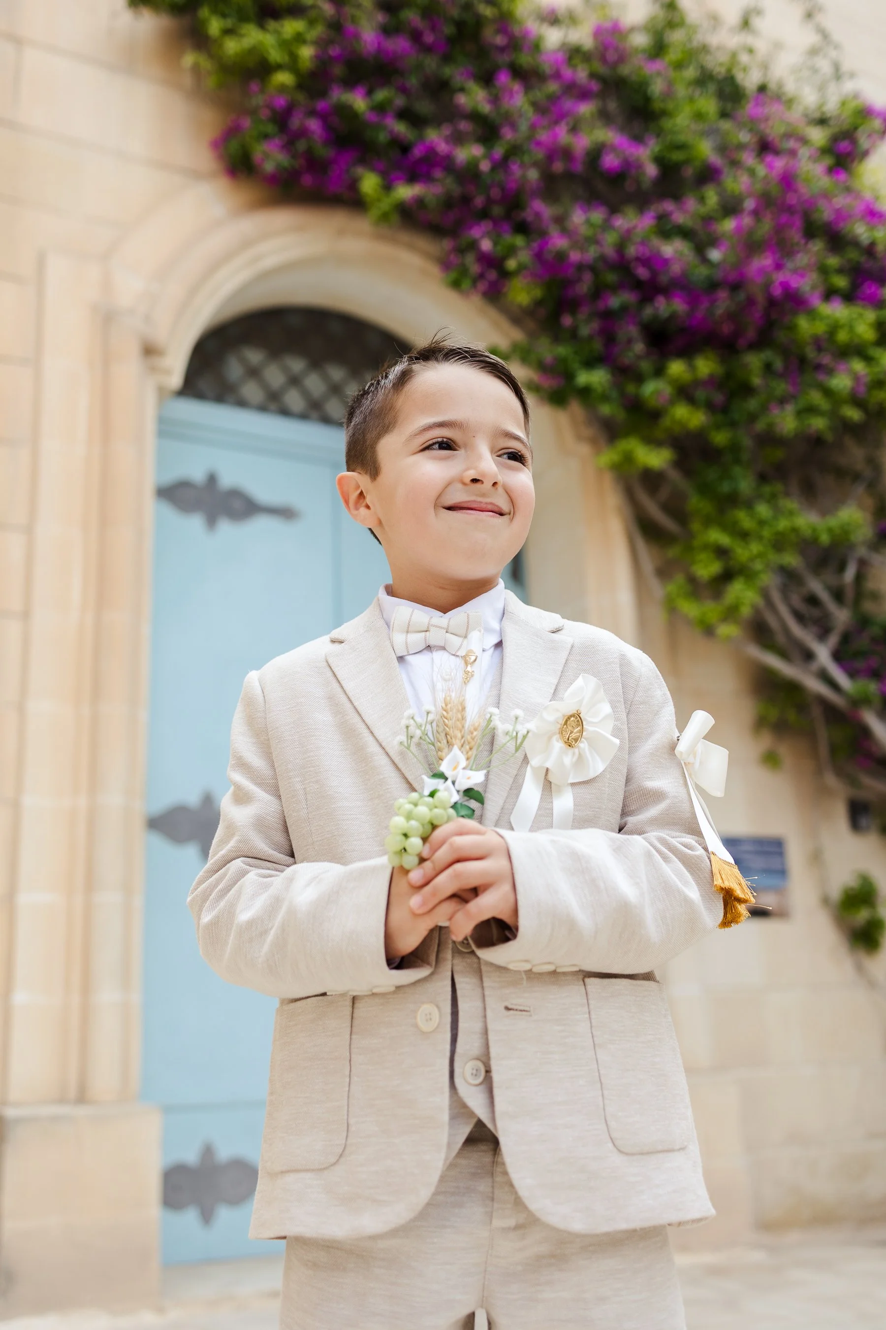 Young boy dressed in a beige suit with a bow tie, holding a small bouquet of flowers, standing in front of a blue door decorated with purple bougainvillea.