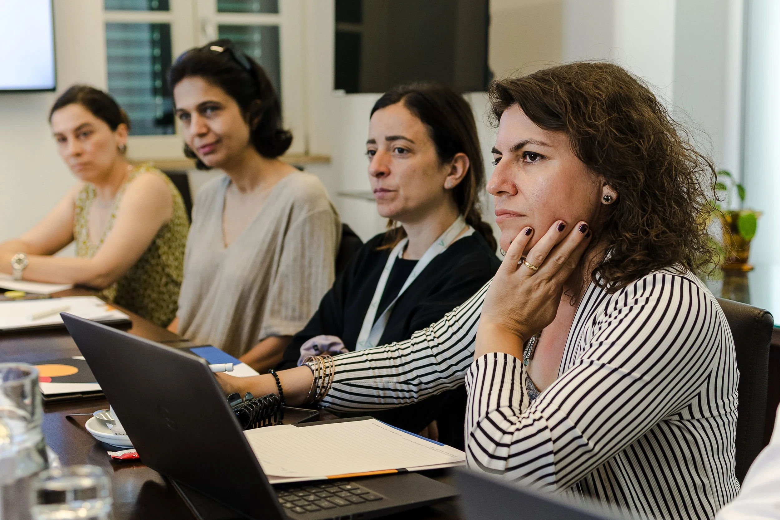 Four women sitting at a conference table during a meeting, with one woman in the foreground wearing a striped blazer and looking attentively.