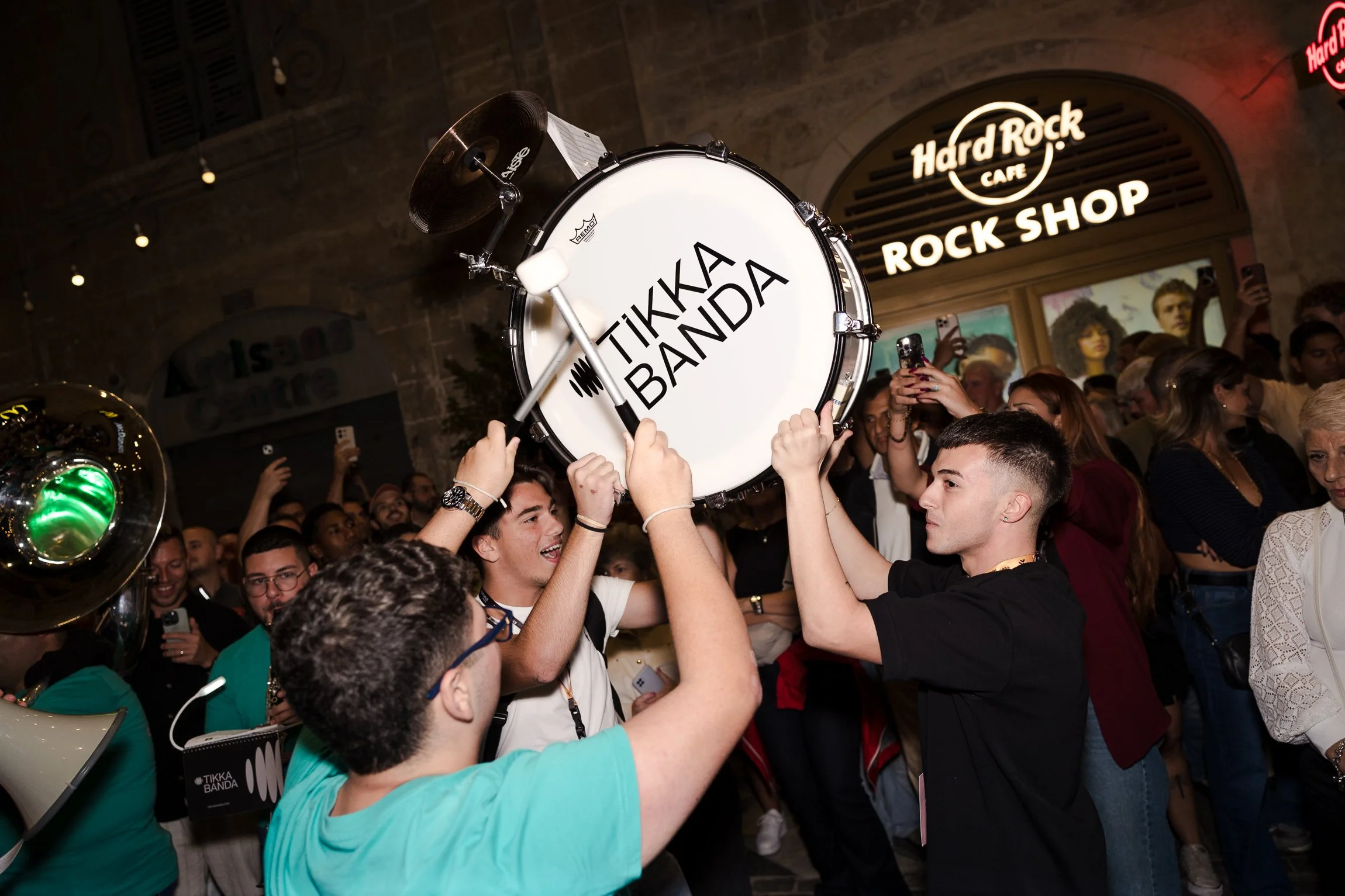A group of people holding a drum with 'TIKKA BAND' written on it, gathered at a concert or event outside a Hard Rock Cafe Rock Shop.