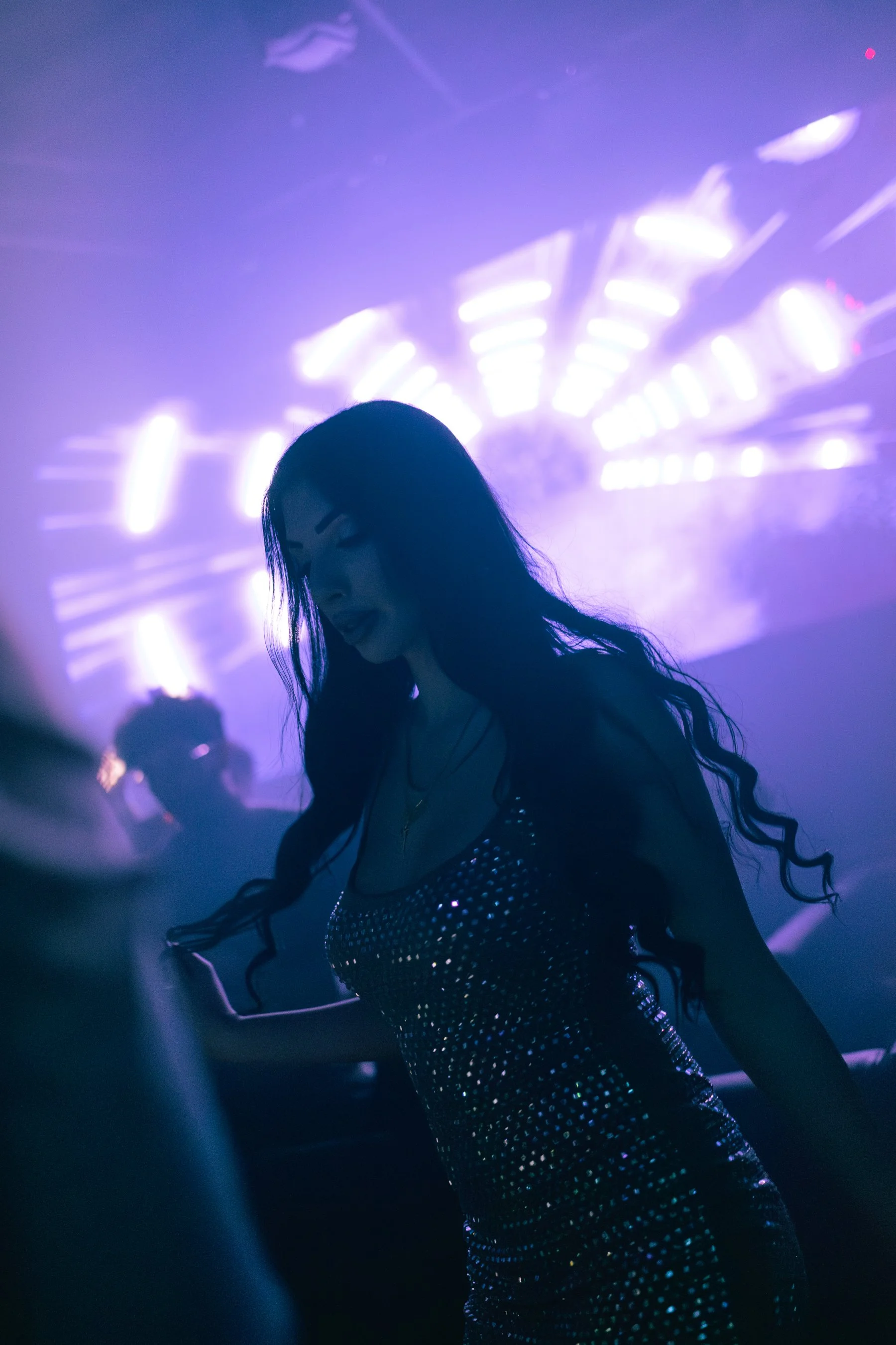 A woman with long dark hair dancing in a club with purple lighting and a light display in the background.