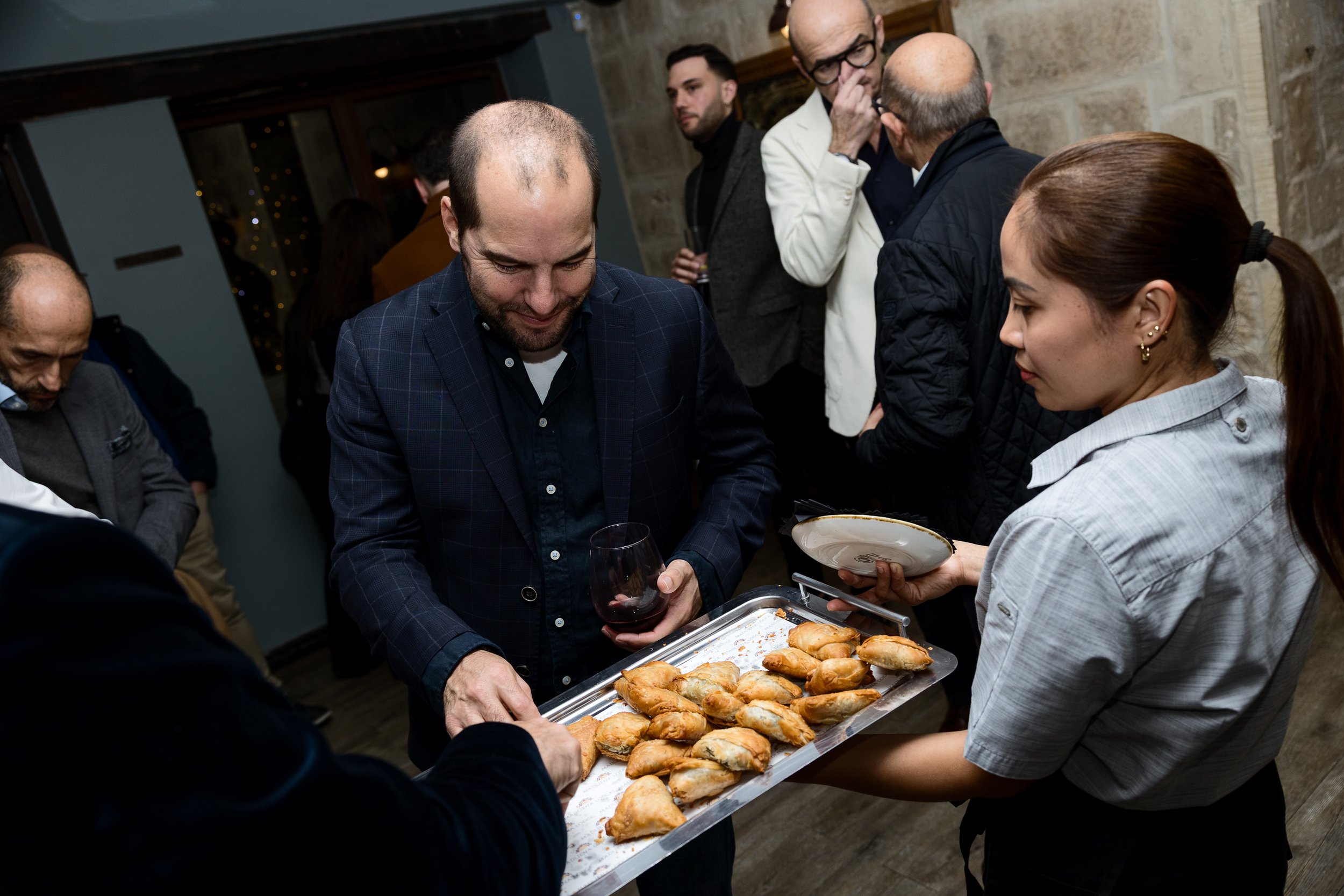 People at a social gathering enjoying finger foods, with a server handing out a tray of small pastries or appetizers.