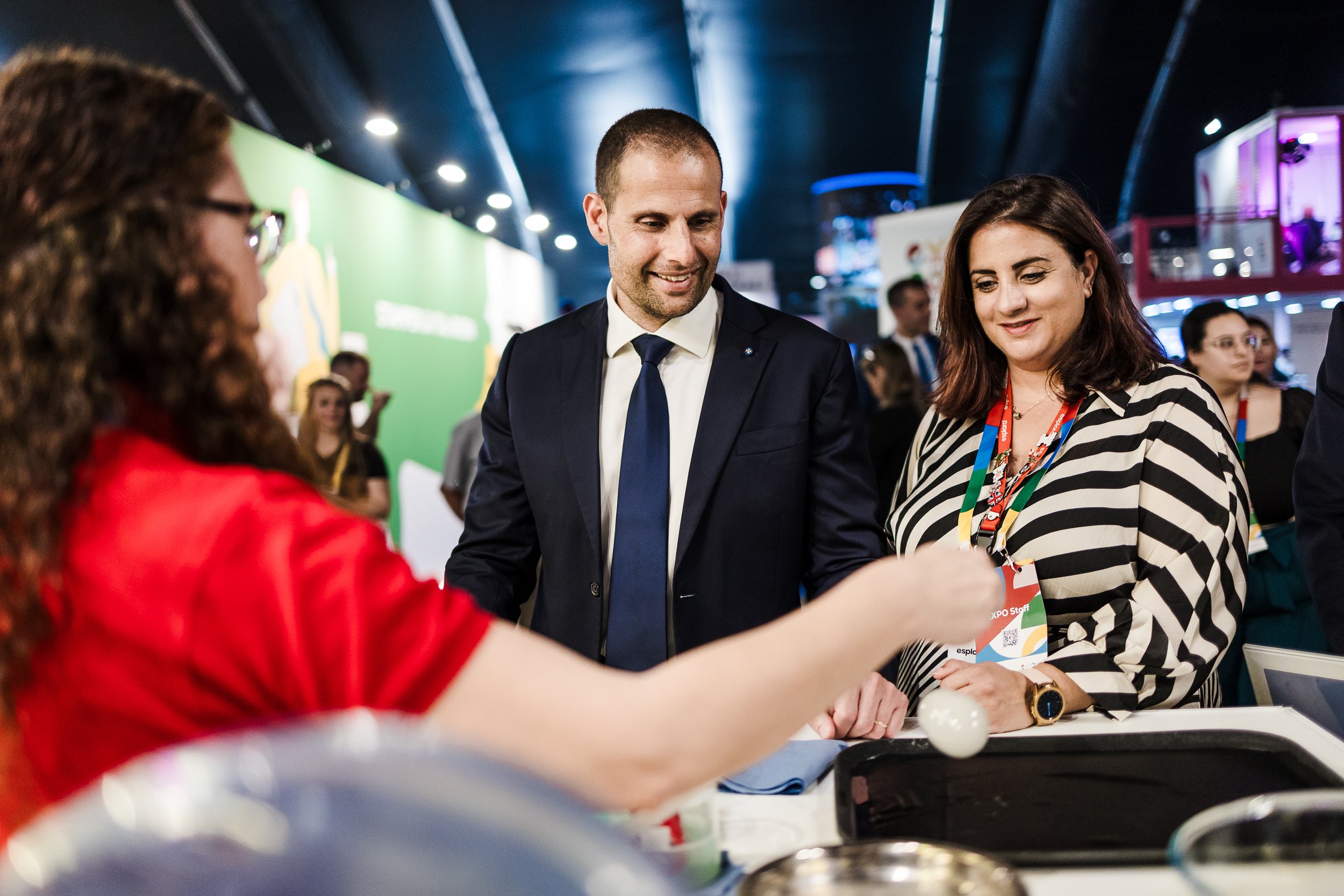 A man in a navy suit and a woman in a black and white striped shirt are smiling and looking at a woman in a red shirt who is demonstrating something at a table, at a busy event or conference.