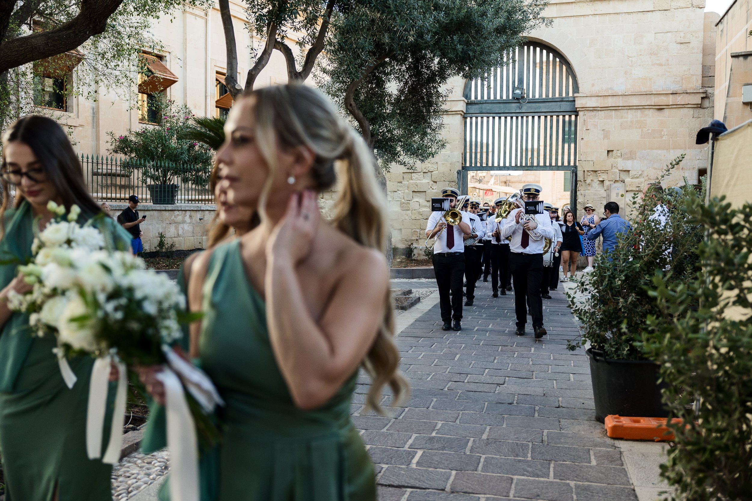 A wedding procession walking outside, with women in green dresses holding bouquets and a marching band playing instruments in the background near a stone wall and trees.