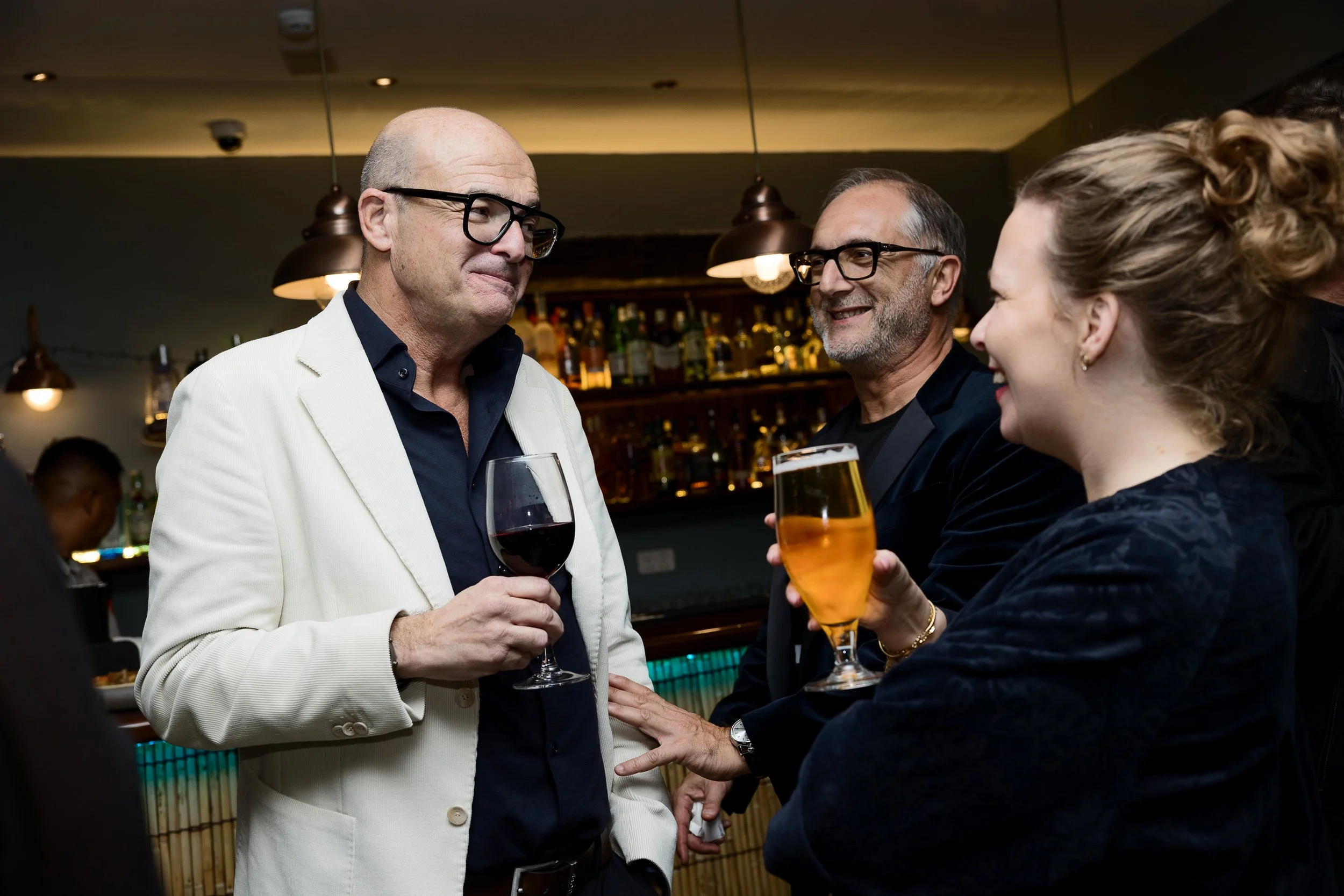 Three people socializing at a bar, two of them holding drinks, a man with glasses in a white jacket, another man with glasses in dark attire, and a woman with curly hair smiling.