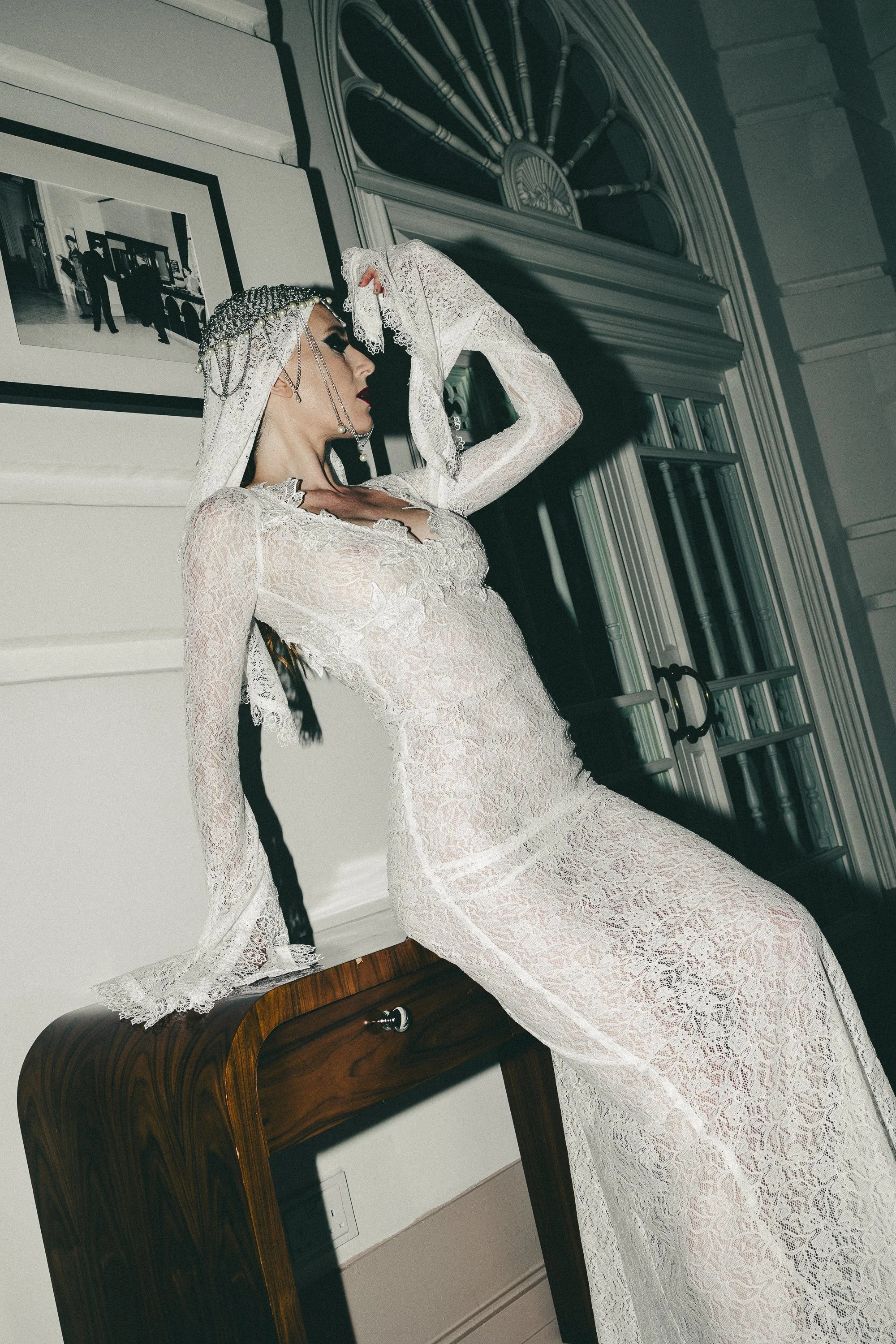 A woman dressed in a white lace gown with long sleeves is posing indoors against a decorative wall and mirror. She is wearing a headpiece with chains, her eyes are closed, and she has striking makeup.
