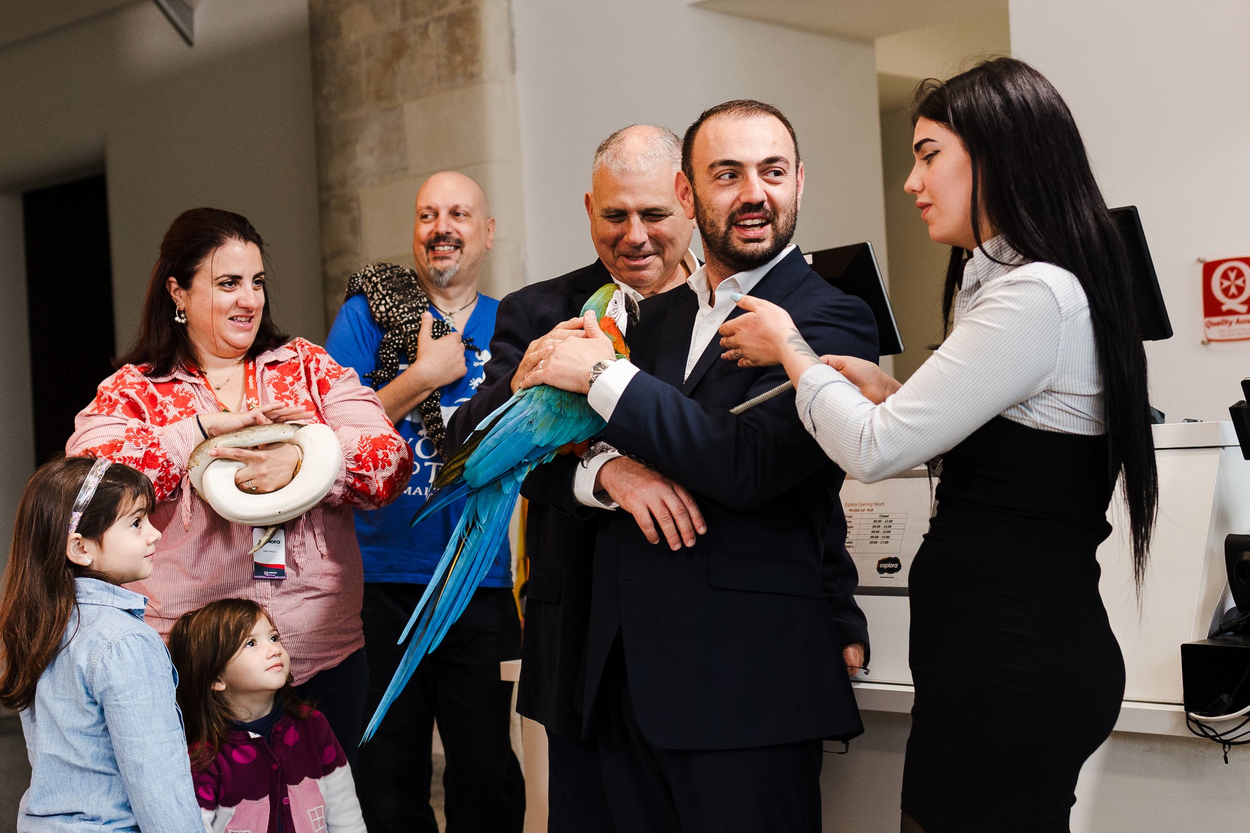 Group of people holding and interacting with a colorful parrot at an indoor event, including two young girls watching.