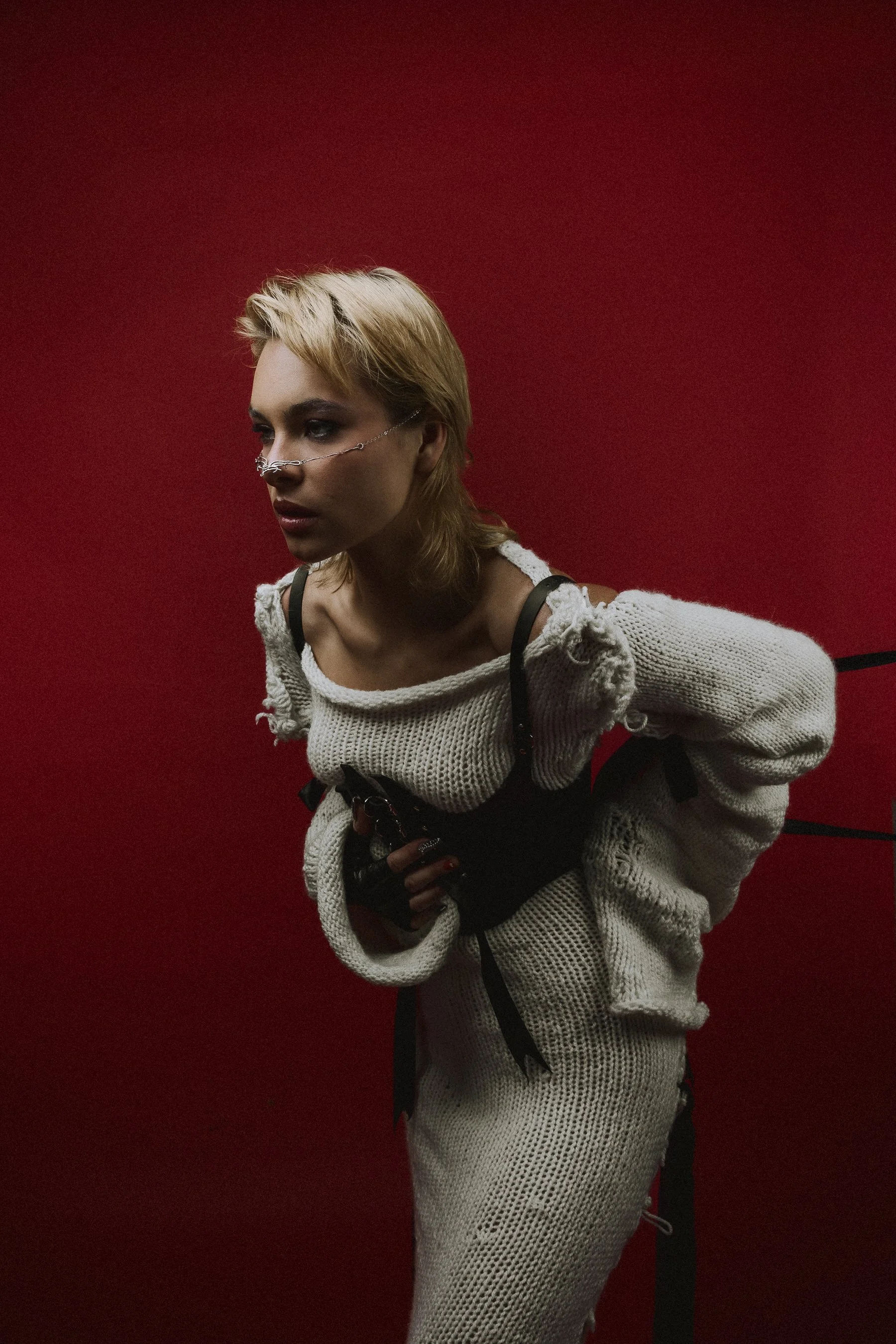 A woman with blonde hair in a messy style, wearing a white chunky knit sweater with distressed details, black gloves, and a black harness, standing against a red background, with a slightly tilted posture and a serious expression.